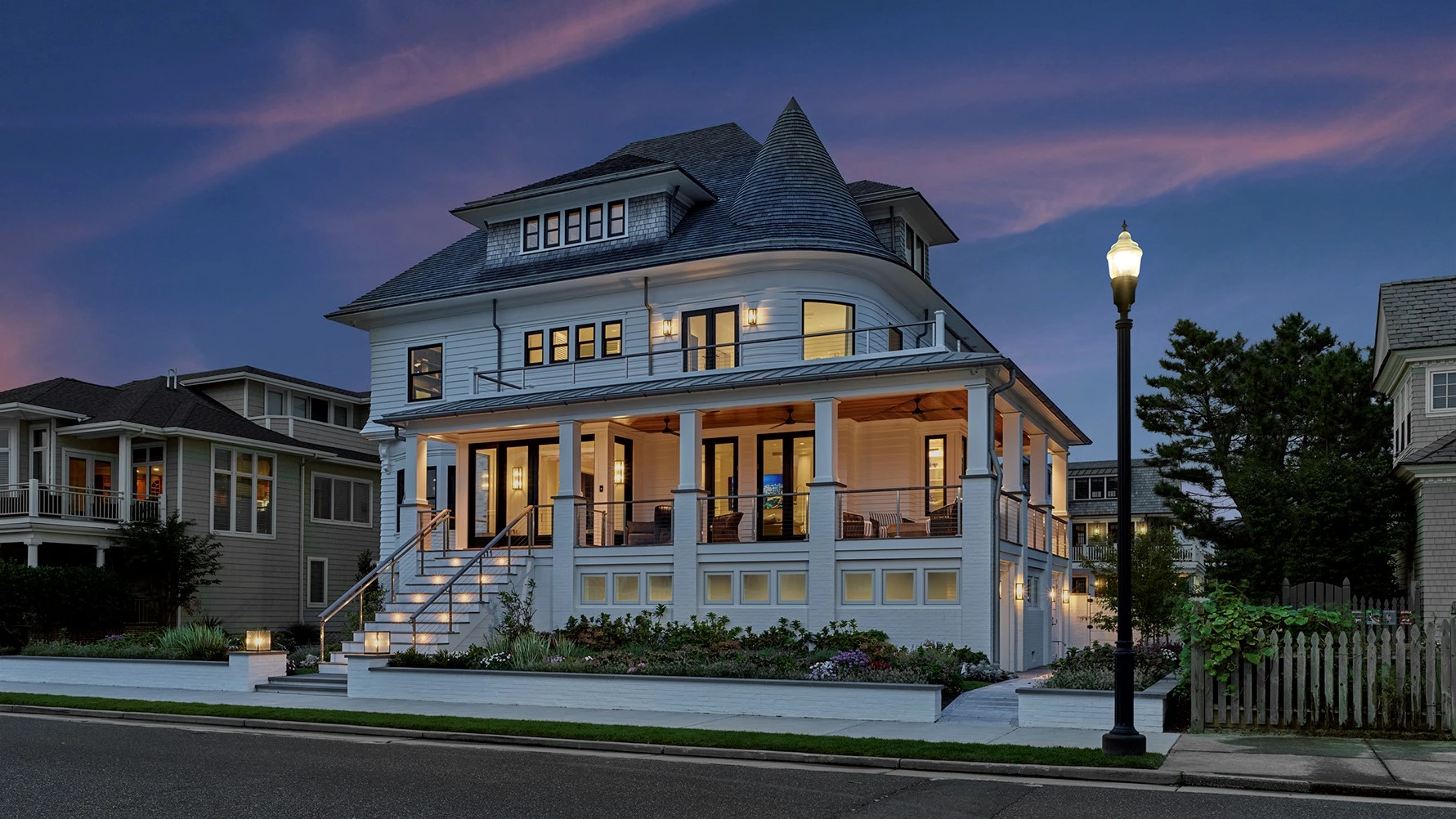 Light blue Victorian-style house with wraparound porch, turret, and illuminated windows at dusk.