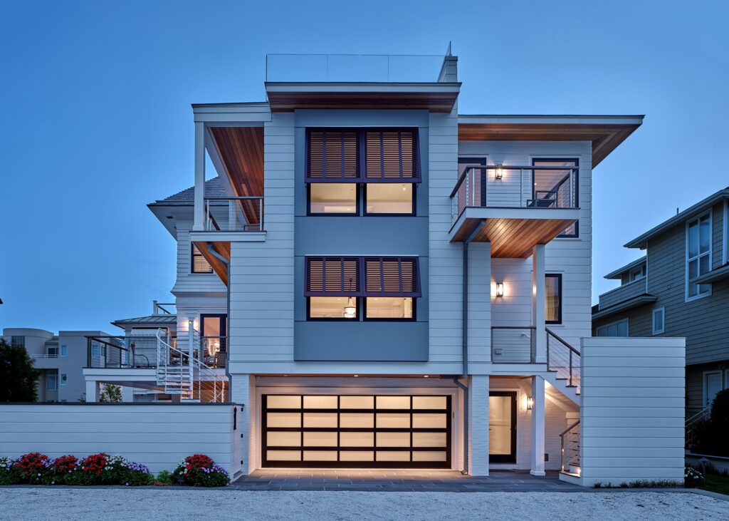 Modern three-story residential house with white horizontal siding, black-framed windows, wood accents, and glass garage doors at dusk.
