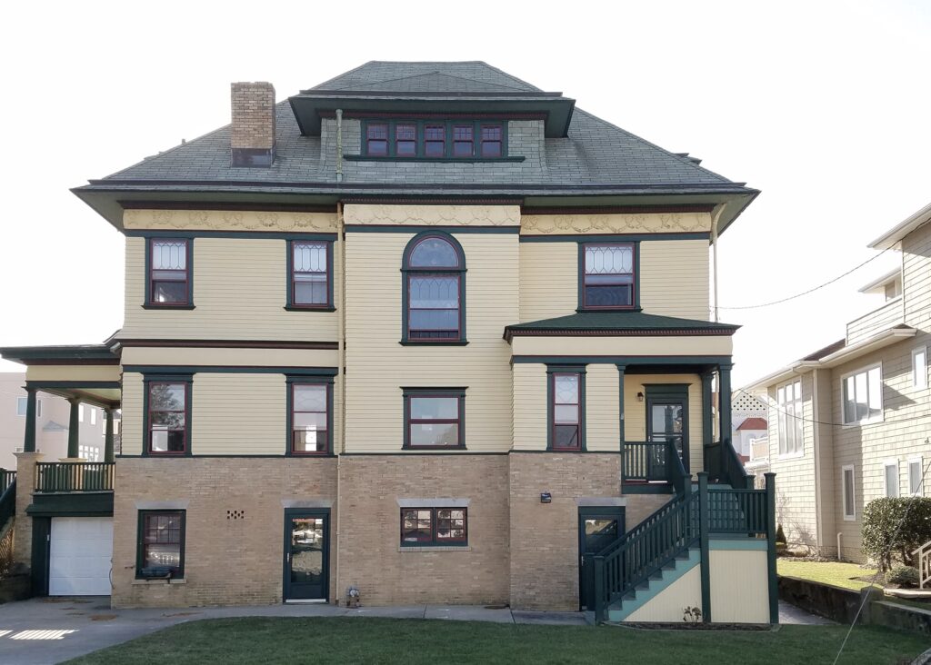 Three-story Victorian mansion with cream clapboard siding, brick base, mansard roof with dormer windows, and dark trim framing arched and rectangular fenestration.
