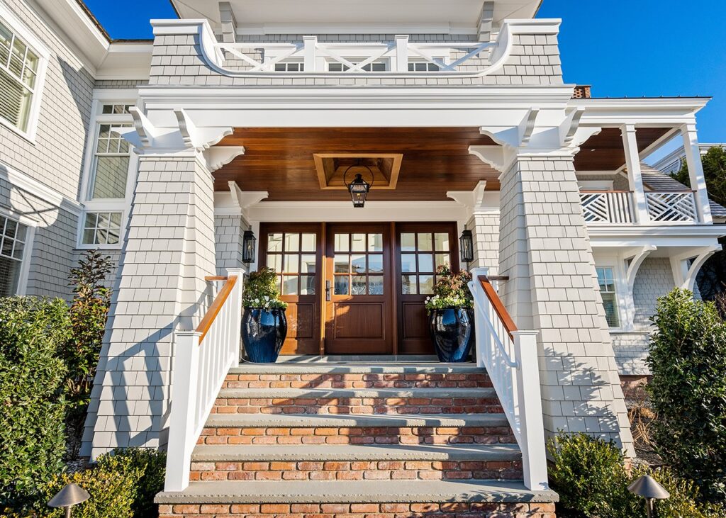Modern coastal home facade featuring shingle siding, white trim, wood soffit detail, and dark wood entry doors with transom windows.