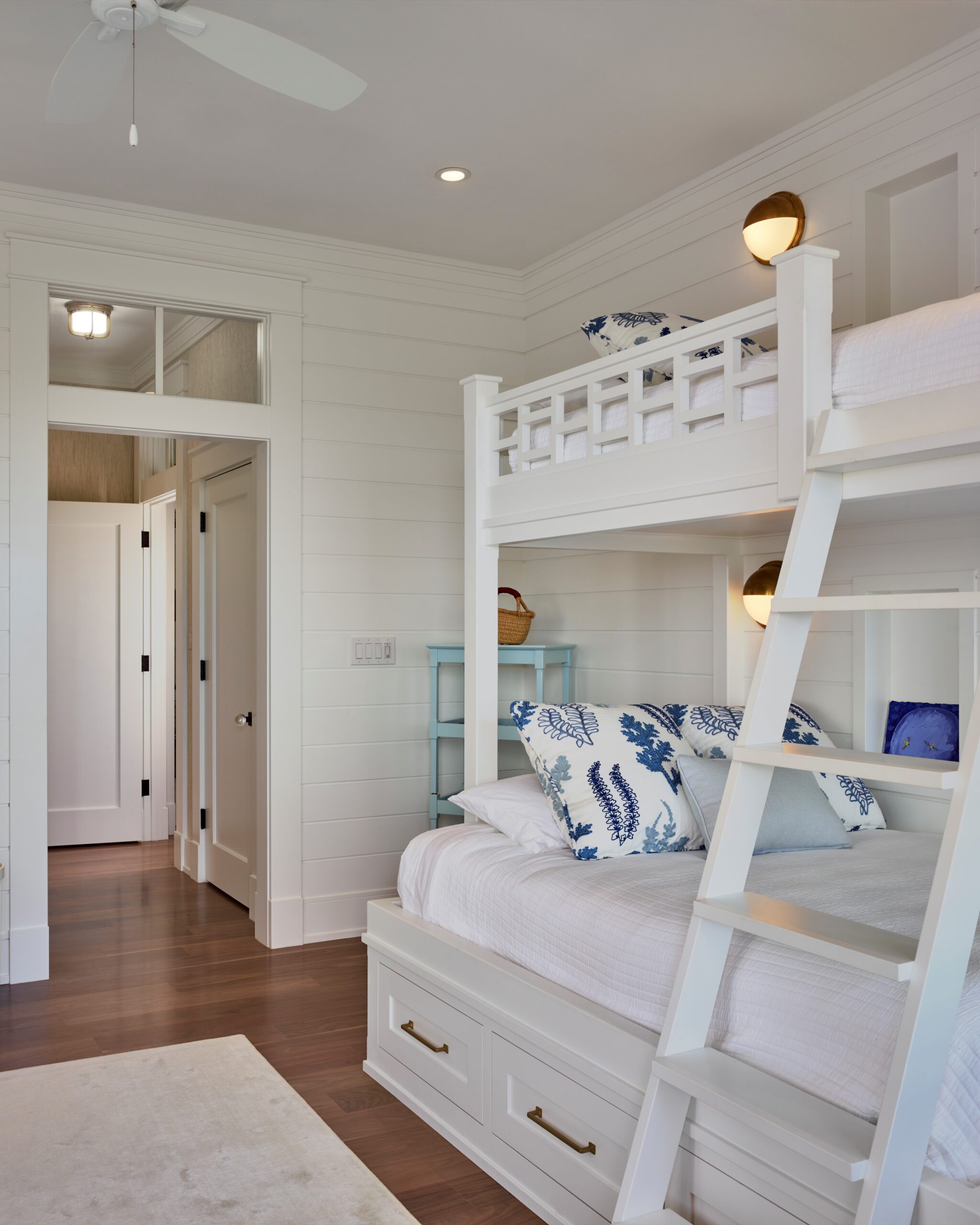 White built-in bunk beds with storage drawers, shiplap walls, and brass sconces in a bright bedroom with hardwood flooring.