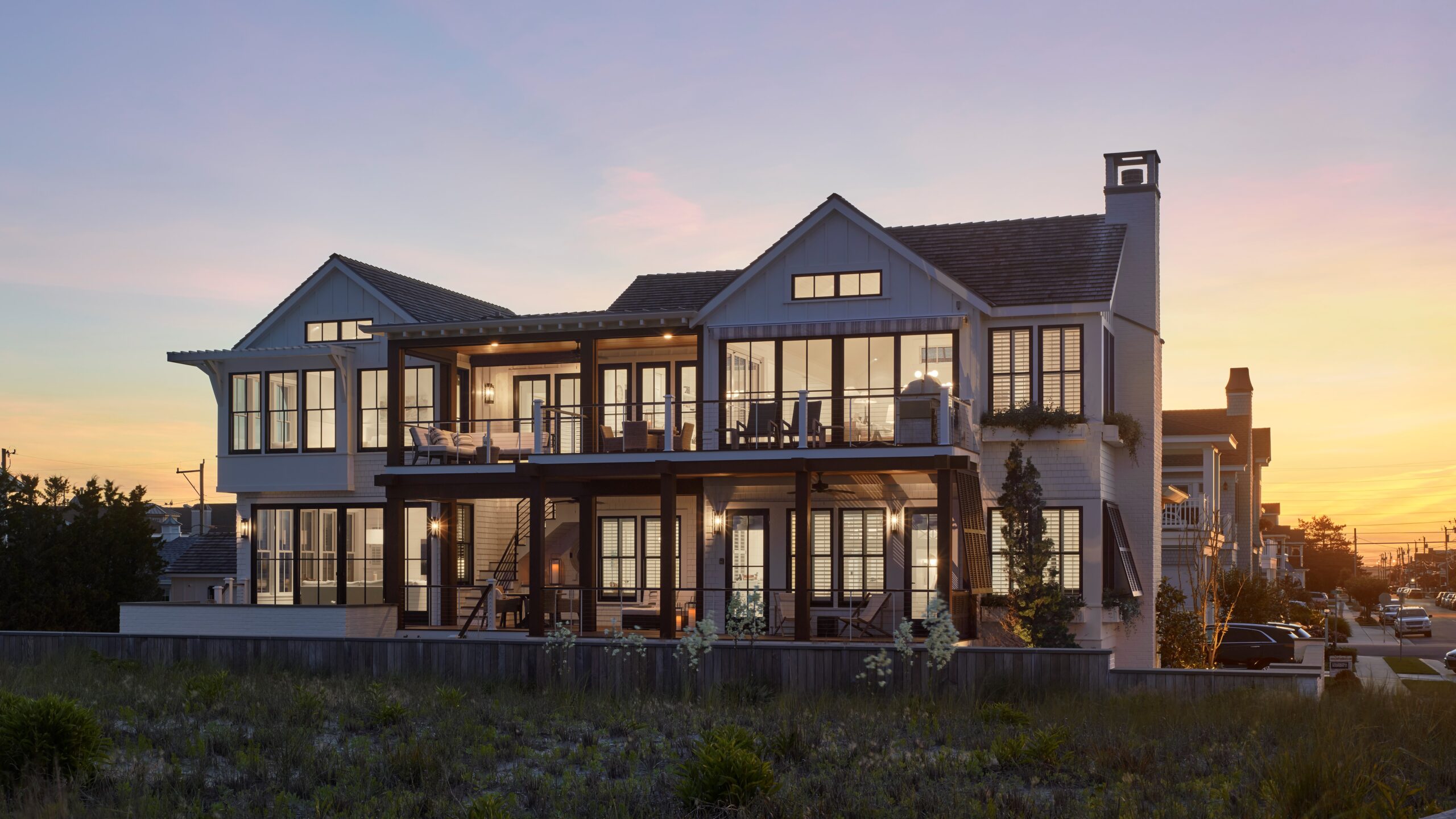 Modern two-story residential home with dark gabled roofline, extensive glazing, and raised deck at dusk.
