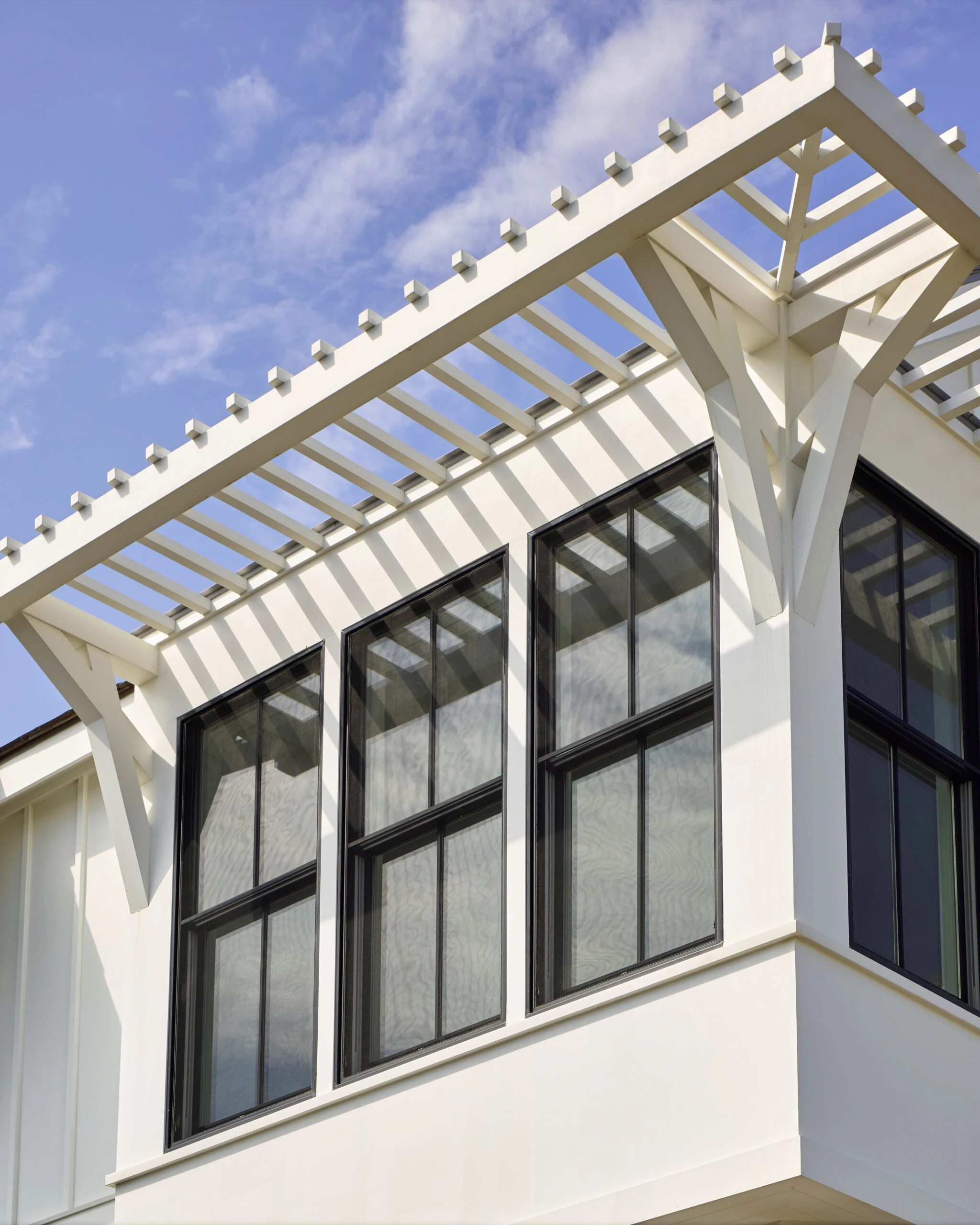 White modern facade with black-framed windows beneath a white pergola structure against blue sky.