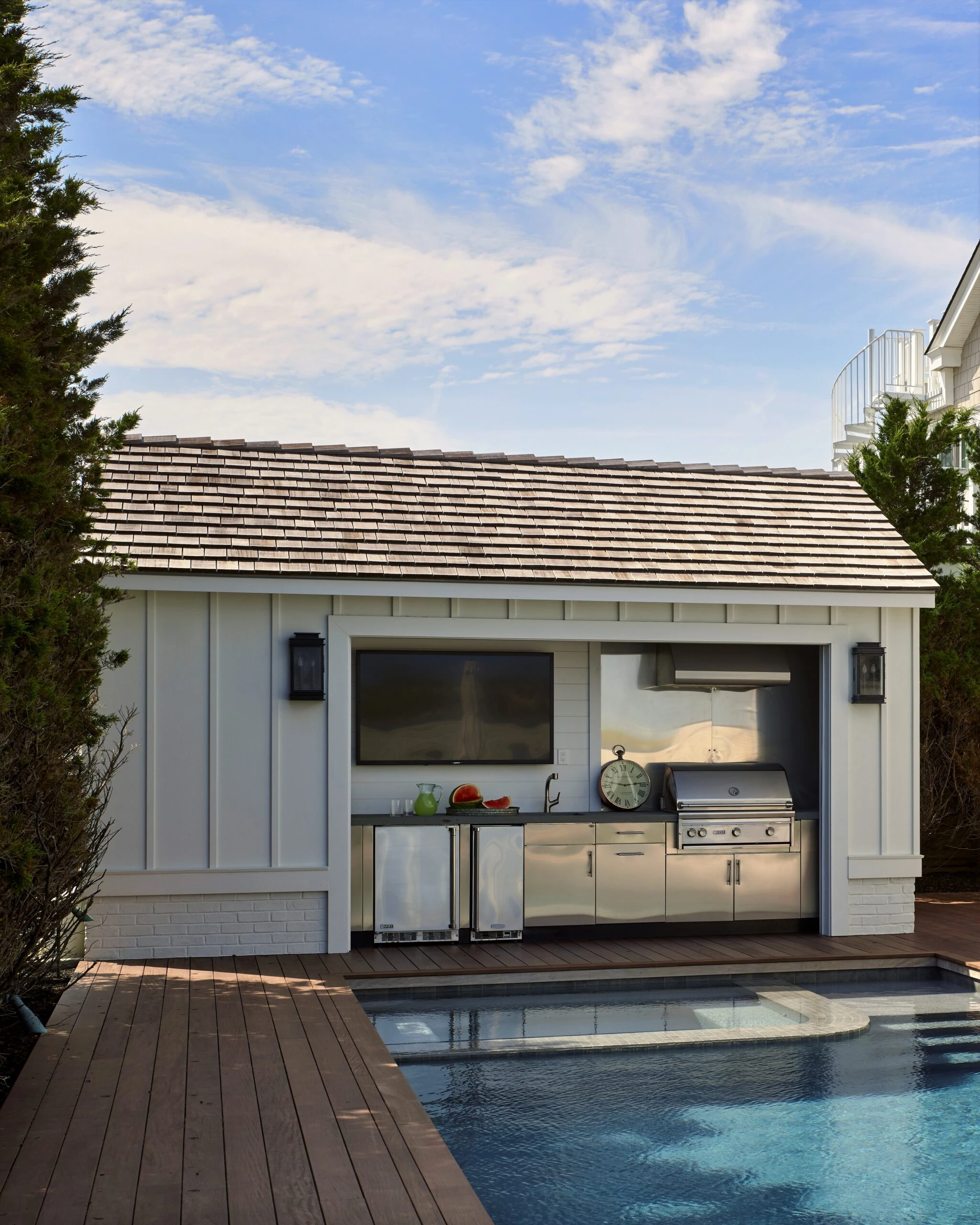 Modern poolside cabana with white board-and-batten siding, slate roof, and integrated stainless steel outdoor kitchen.