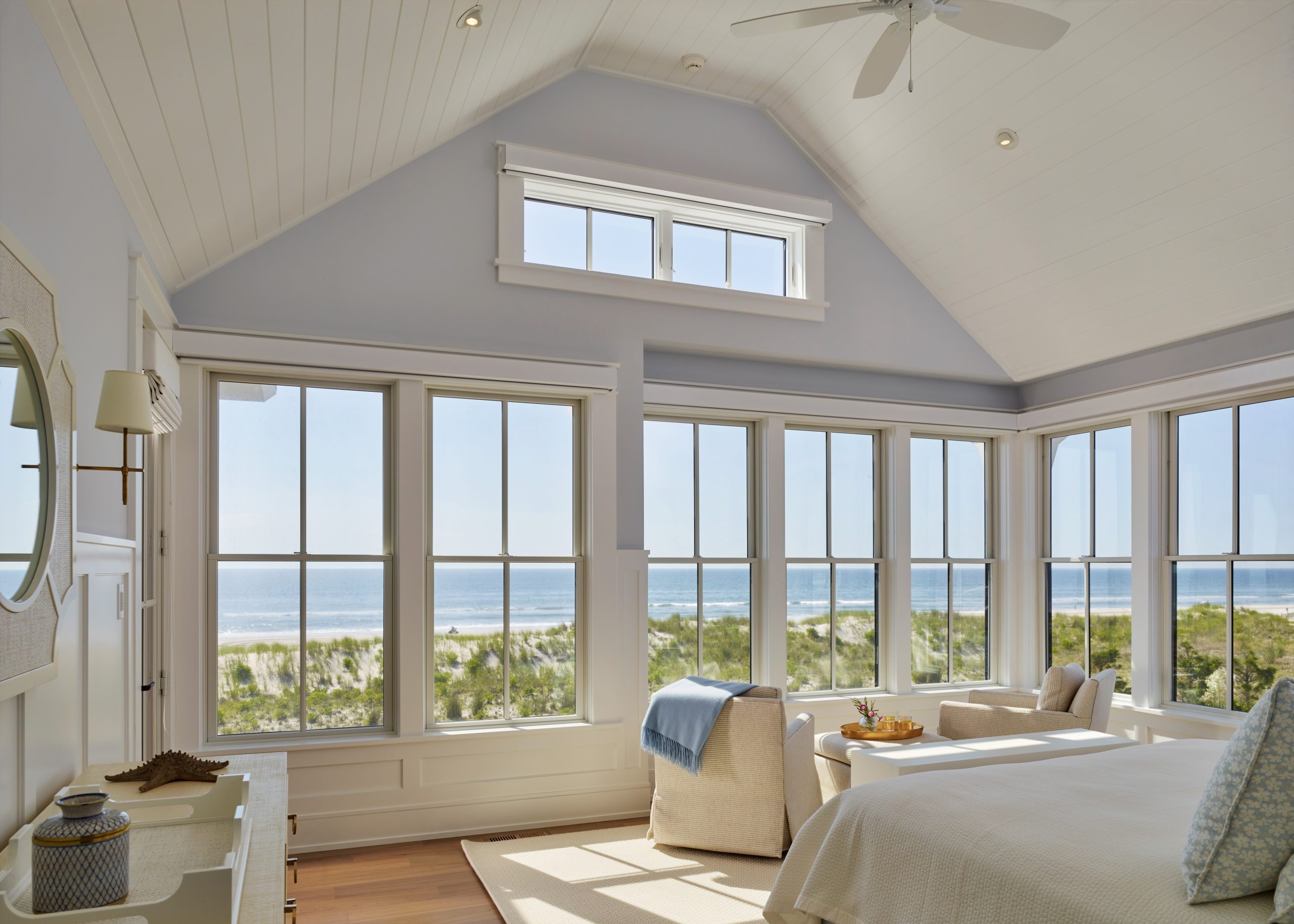 Bright coastal bedroom with vaulted ceiling, clerestory window, and expansive multi-pane casement windows overlooking beach and dunes.