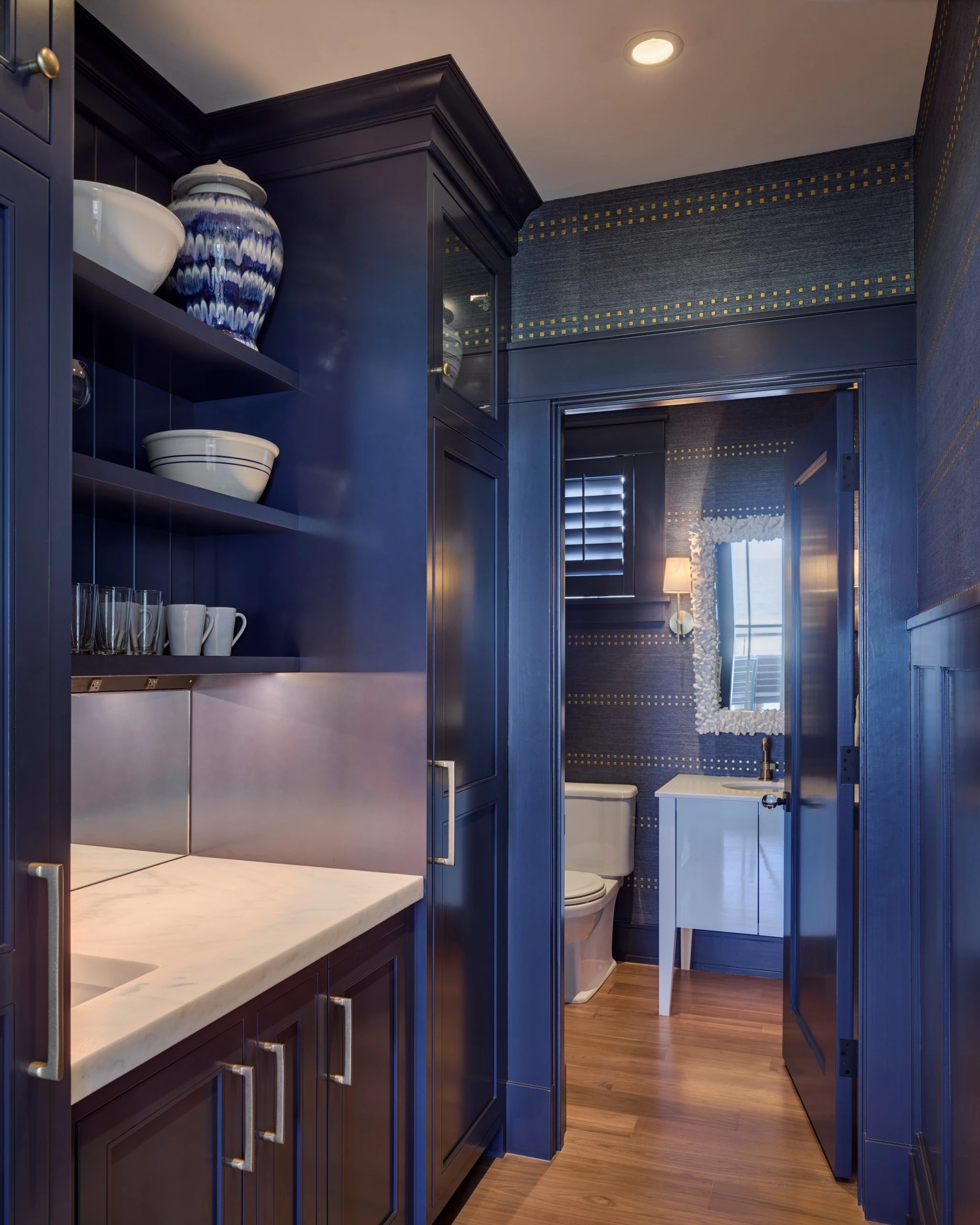 Navy blue bathroom with brass-studded wallpaper, dark cabinetry, white quartz countertops, and glass-enclosed shower beyond doorway.