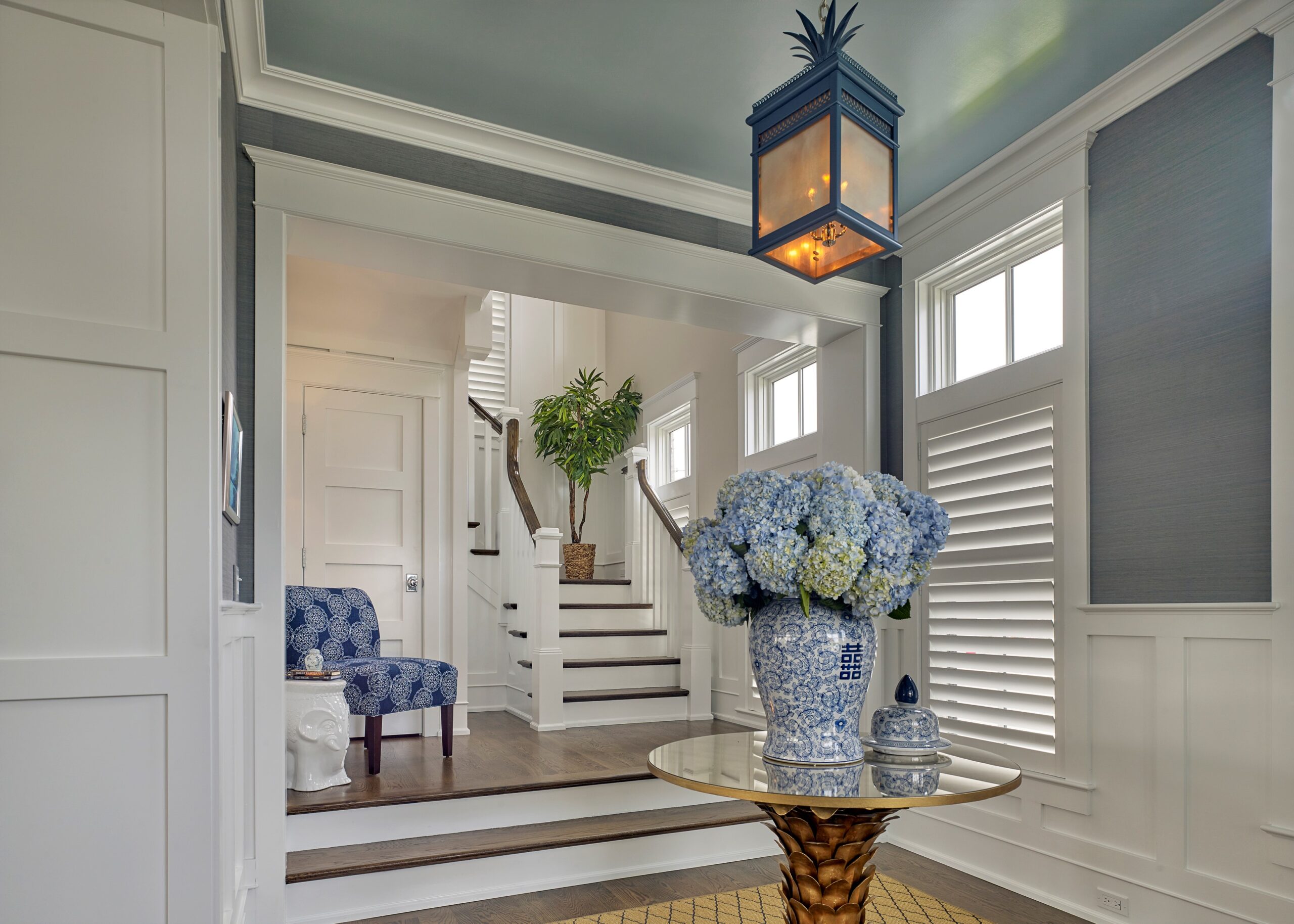 Foyer with gray walls, white trim, pendant lantern, brass pedestal table with blue-and-white porcelain, and staircase visible beyond.