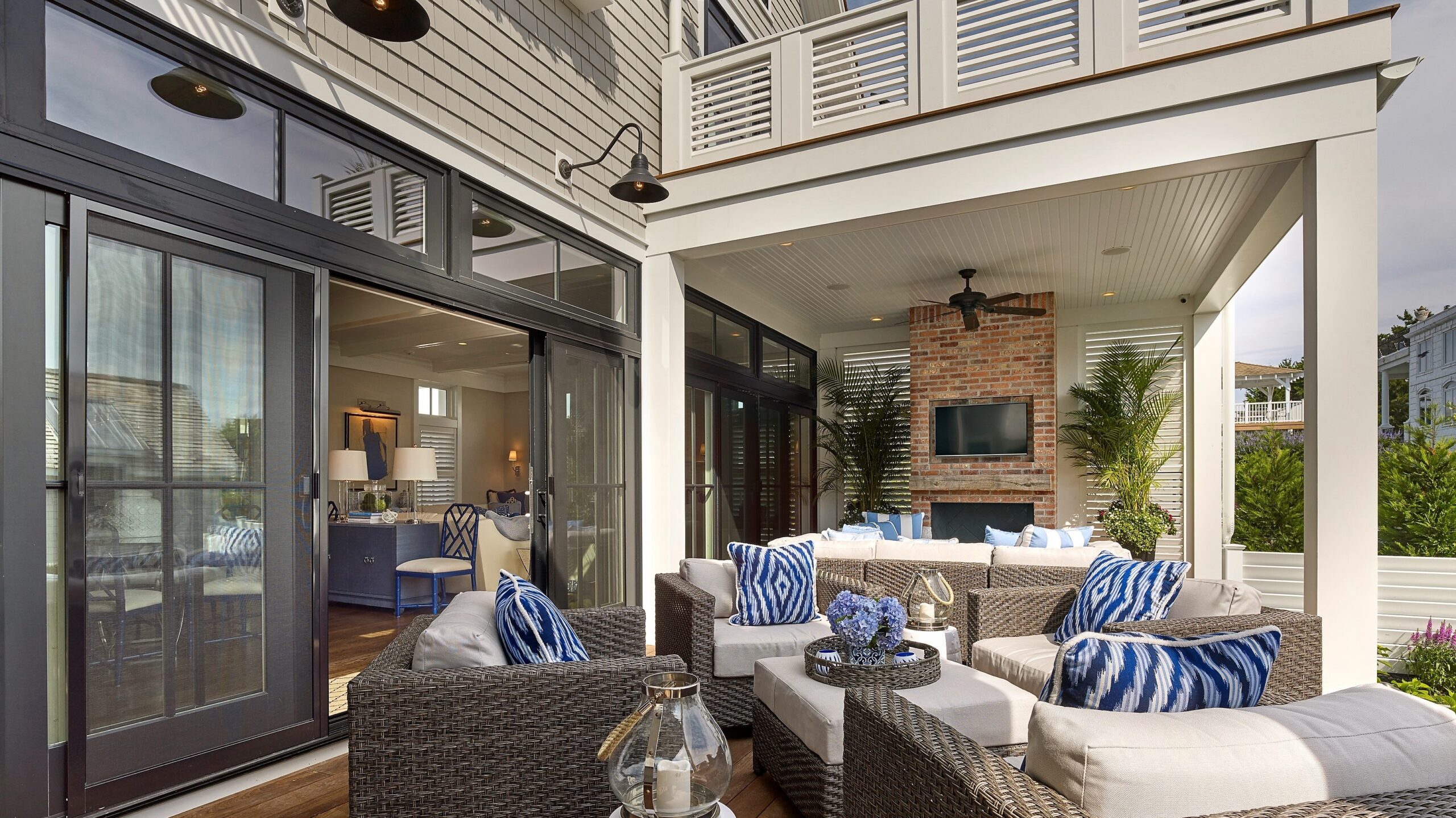 Modern covered patio with woven furniture, brick fireplace, clerestory windows, and dark-framed sliding glass doors opening to interior spaces.