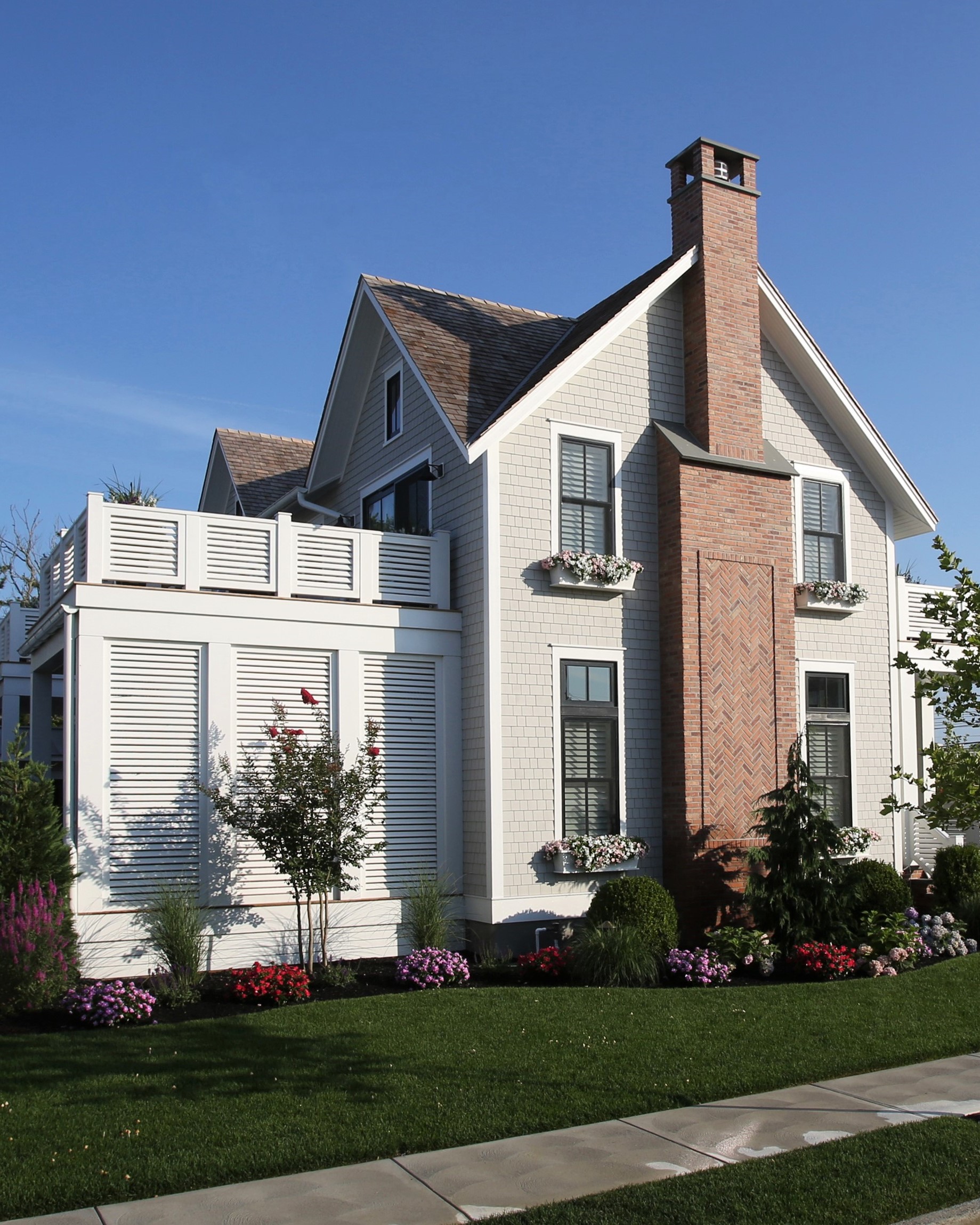 Modern cottage-style home featuring white shingle siding, red brick chimney, and horizontal louvered privacy screen on the street-facing elevation.