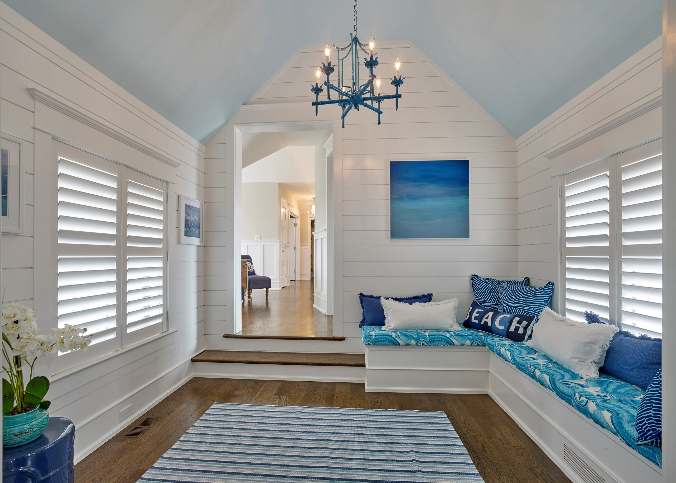 Coastal-style bedroom with shiplap walls, built-in window seat, vaulted ceiling with blue star chandelier, and shuttered windows.