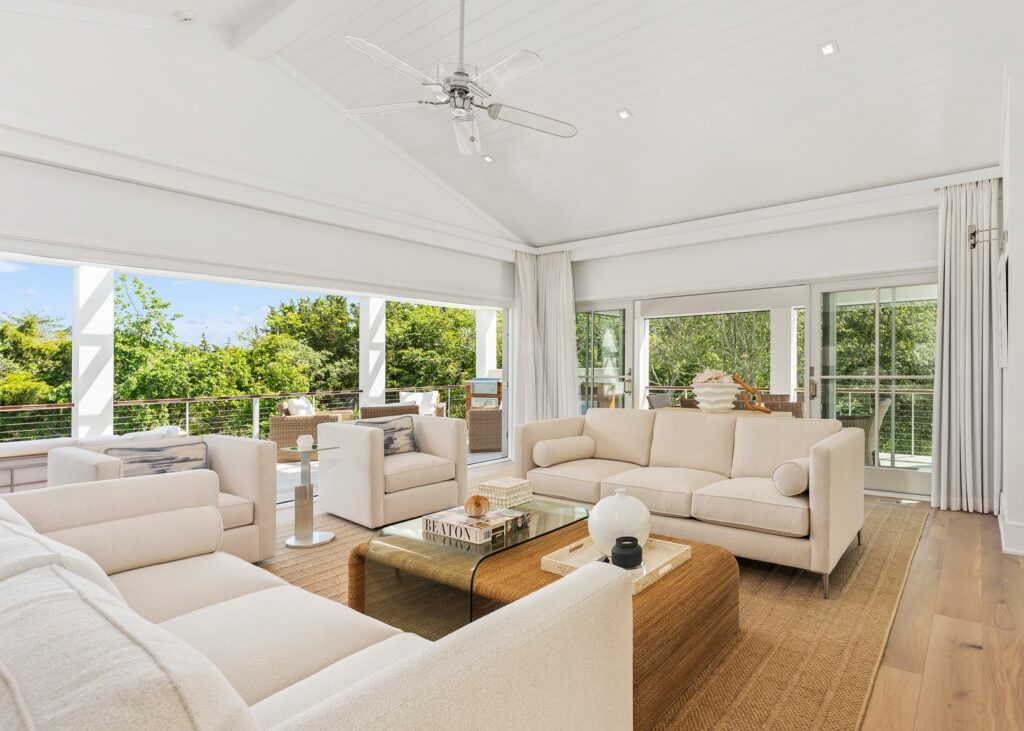 Modern open-plan living room with floor-to-ceiling glazing, white coffered ceiling, ceiling fan, light oak flooring, and cream upholstered furnishings.