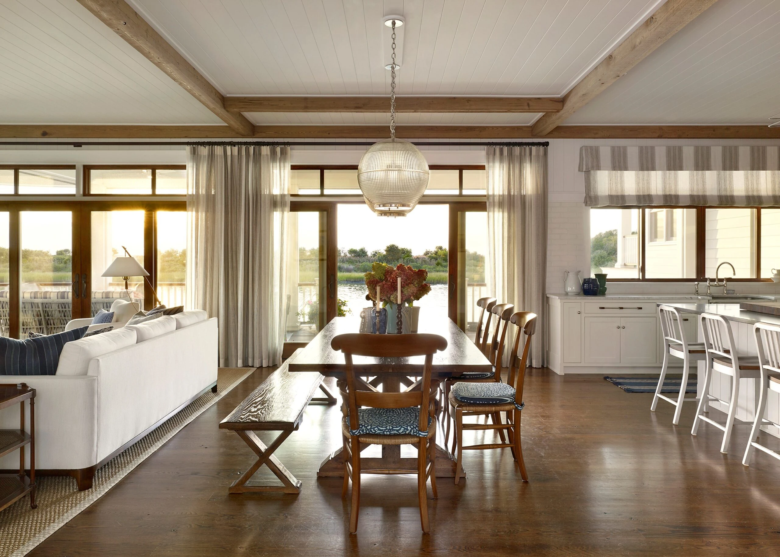 Open-plan dining and kitchen space with exposed wood beams, glazed doors opening to waterfront views, and pendant lighting.