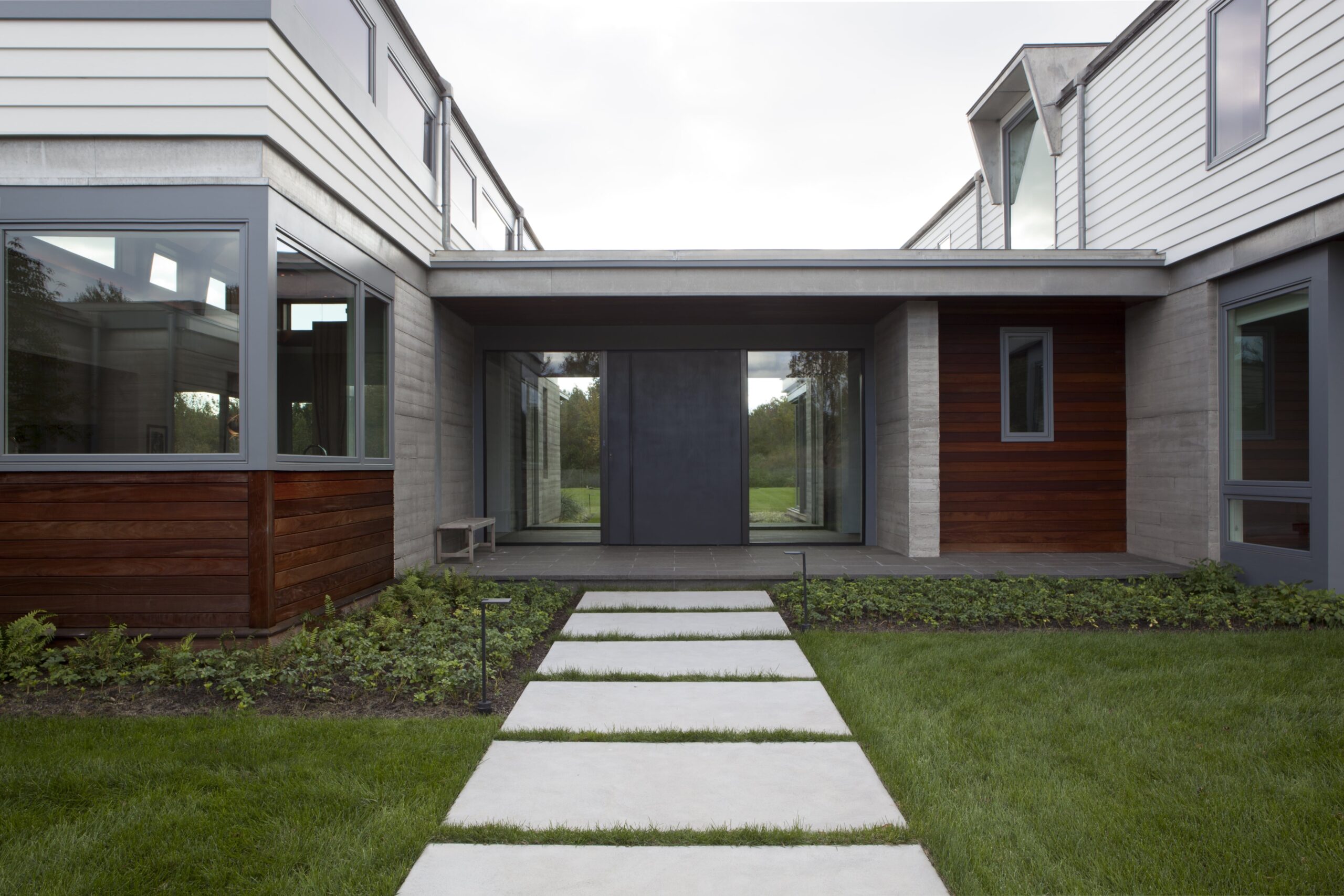 Modern courtyard residence with concrete and wood-clad facades, large glass doors, and linear stepping stone pathway.