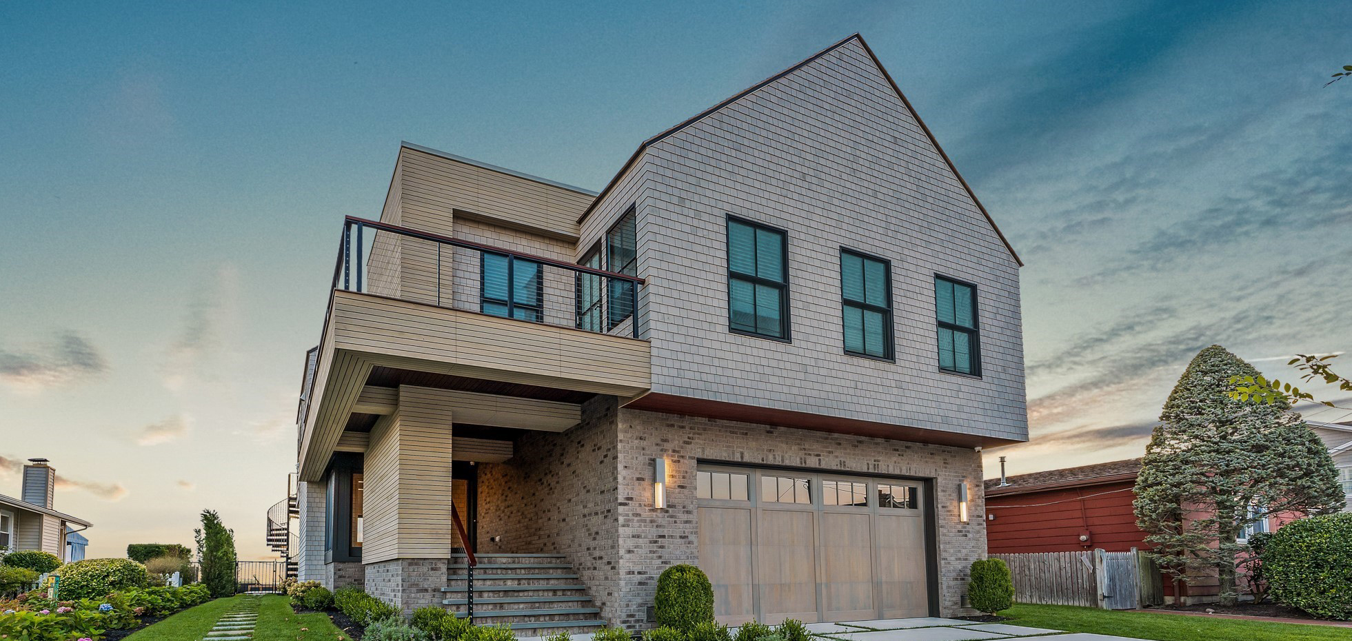 Modern two-story residential home with mixed materials including stone base, horizontal siding, and shingle upper facade with gable roof and metal railings.