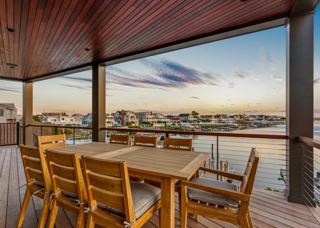 Waterfront deck with wood dining table, horizontal cable railing, wood soffit, and bay view at sunset.