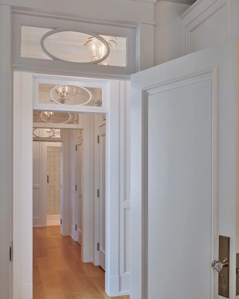 Hallway with white painted trim, transom windows with oval muntins, and wood flooring leading to bathroom.