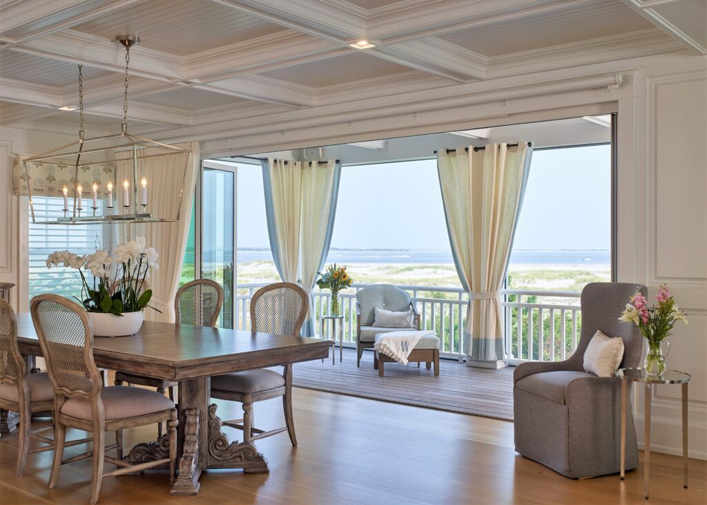 Elegant coastal dining room with coffered ceiling, floor-to-ceiling glass doors, and water views from the railed balcony.