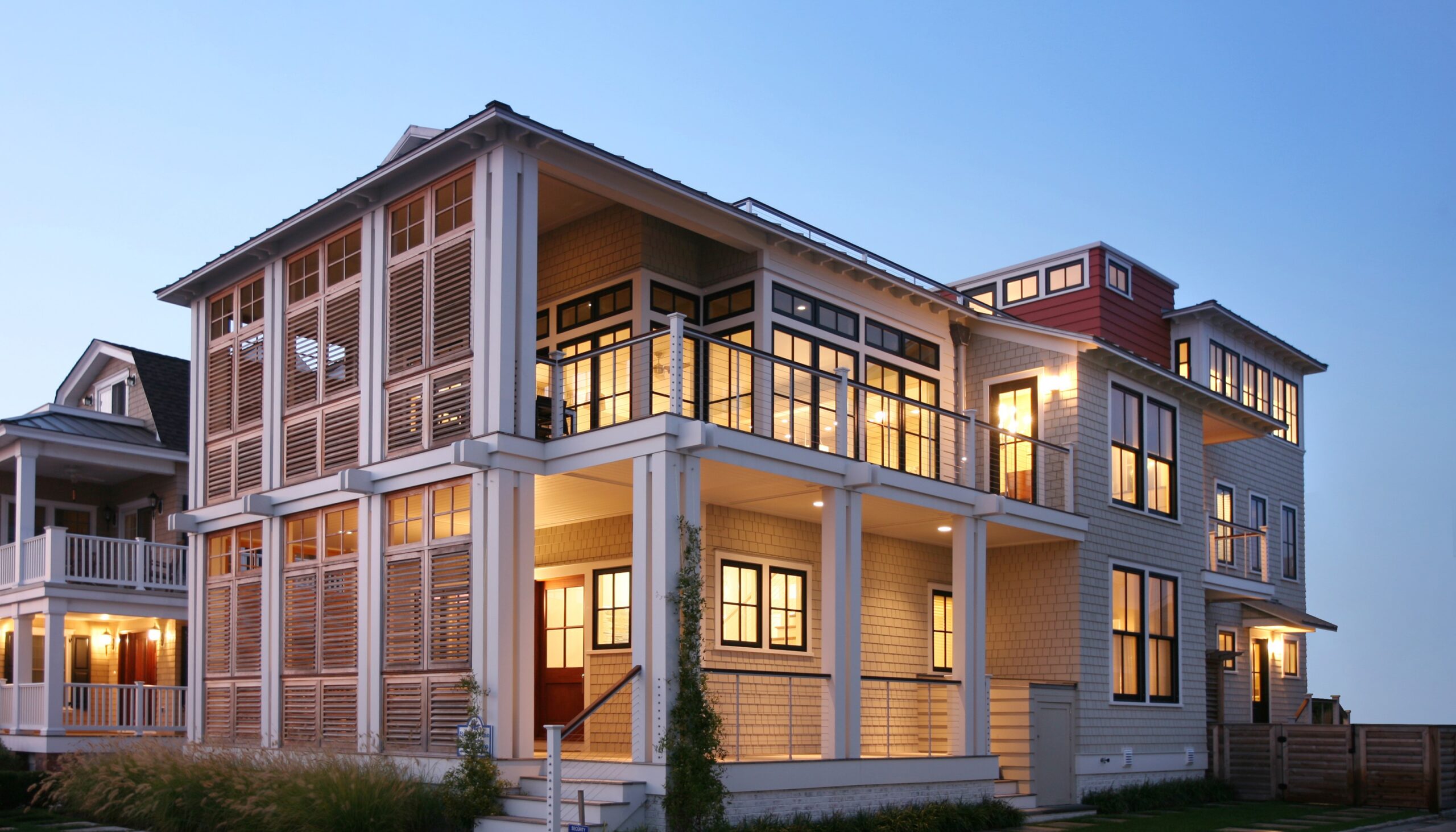Modern coastal residence with shingle siding, multi-level facade, extensive glazing, and horizontal louvered screens at dusk.