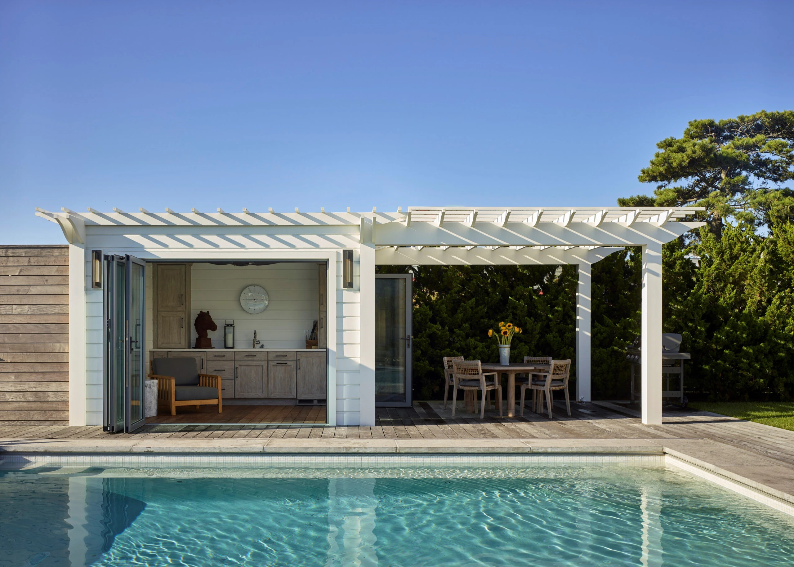 Modern poolhouse with white pergola, sliding glass doors, gray cabinetry, and wooden deck overlooking swimming pool.