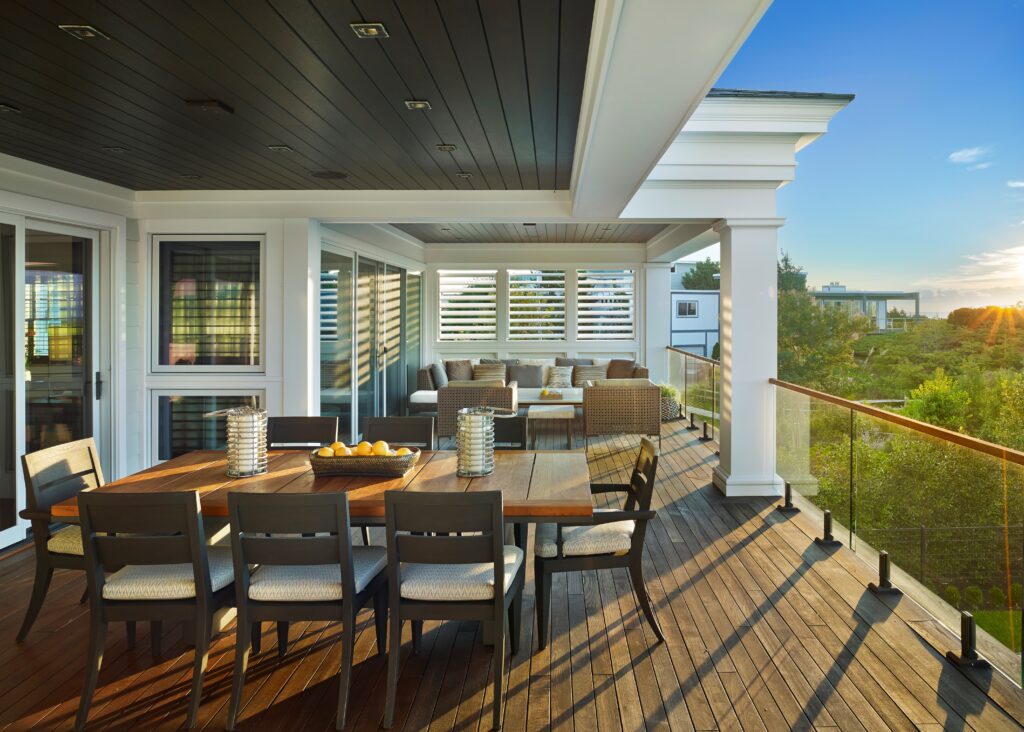 Modern rooftop deck with wooden dining table, louvered shutters, white columns, and cable railing overlooking landscaped grounds.