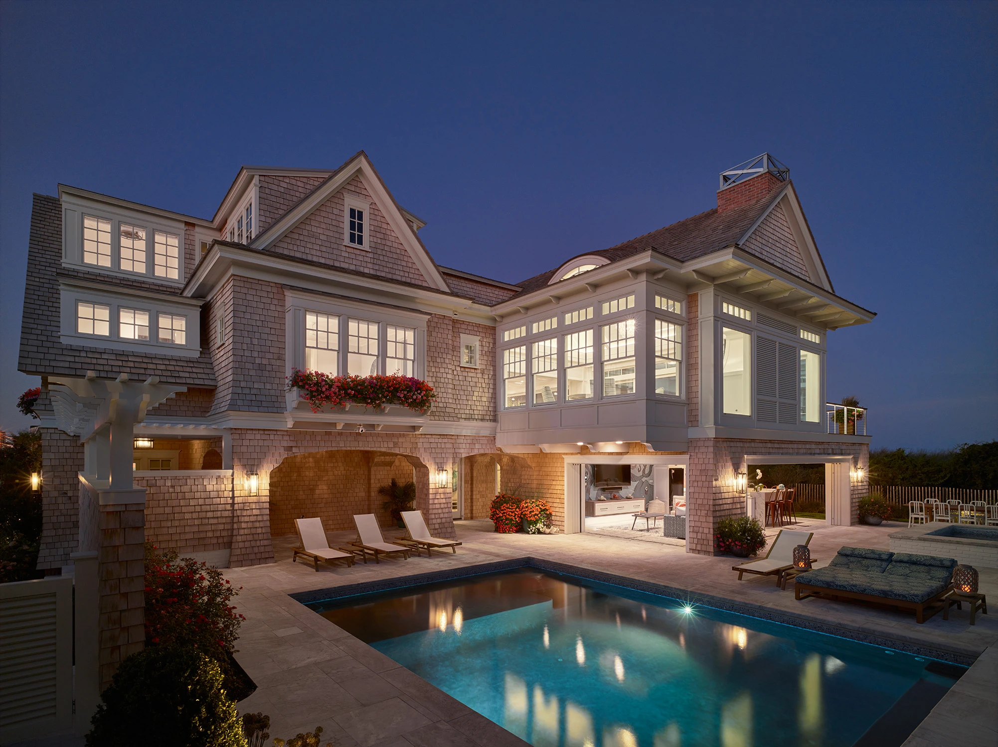 Coastal shingle-clad residence with gabled rooflines, illuminated at dusk, featuring a screened pavilion and pool deck.