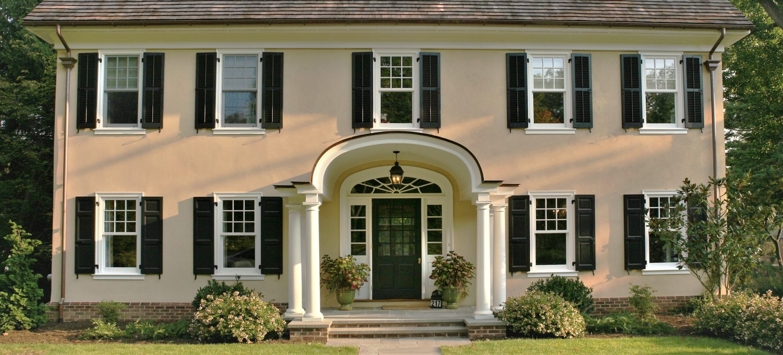Colonial-style residence with cream stucco facade, brick base, arched portico entry, paired columns, and symmetrical window arrangement with black shutters.