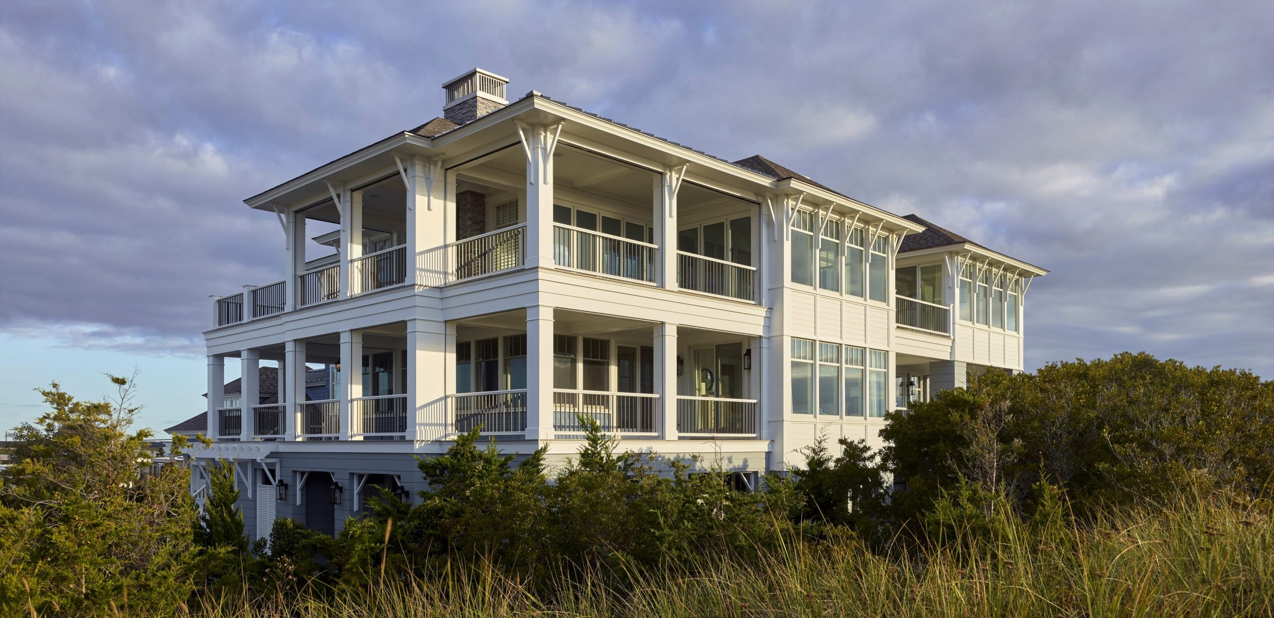 Modern coastal residence with elevated foundation, cream-colored board-and-batten siding, and wraparound porches with metal railings.