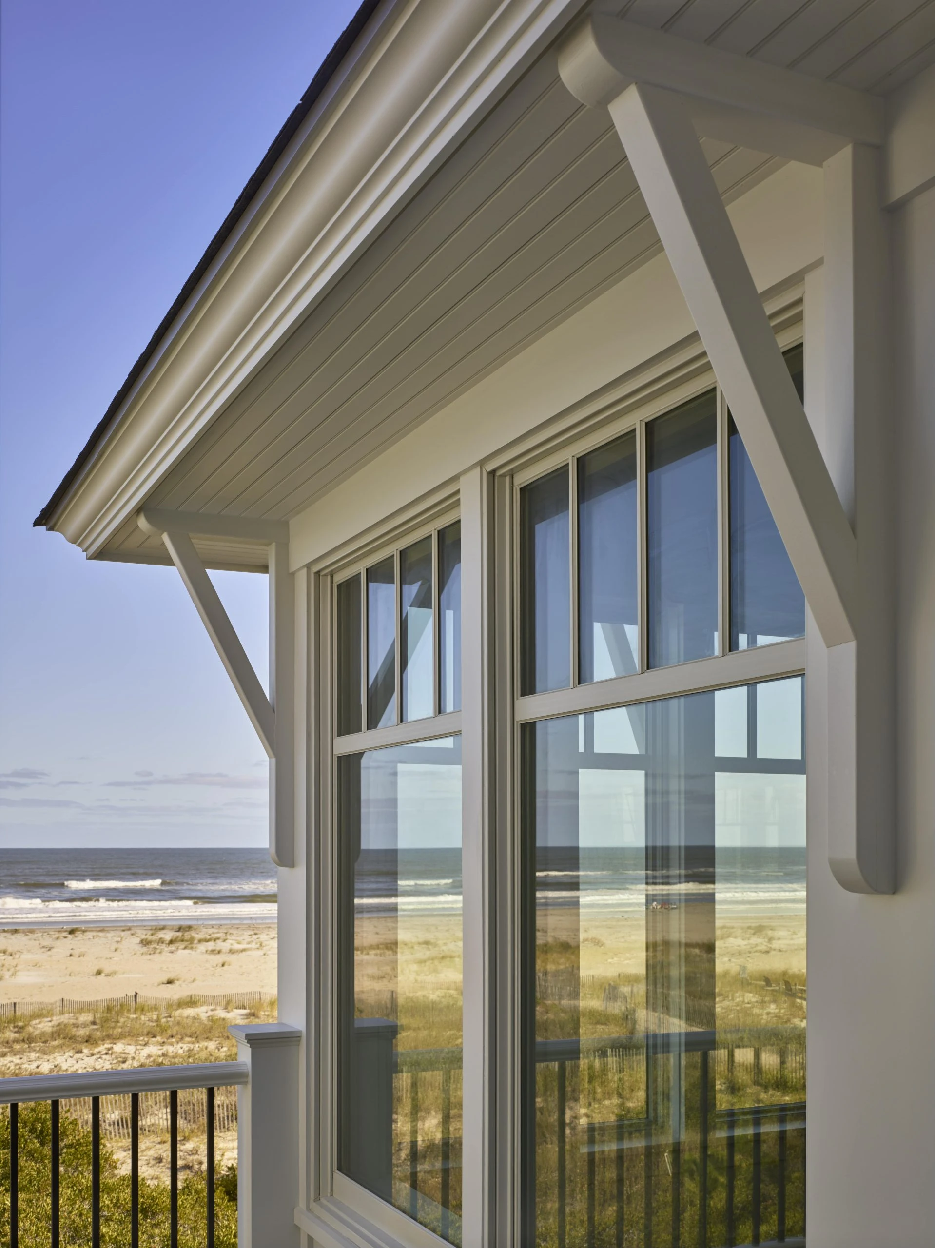 Modern coastal residence with white painted wood siding, multi-pane casement windows, and exposed beam structural elements overlooking beach landscape.