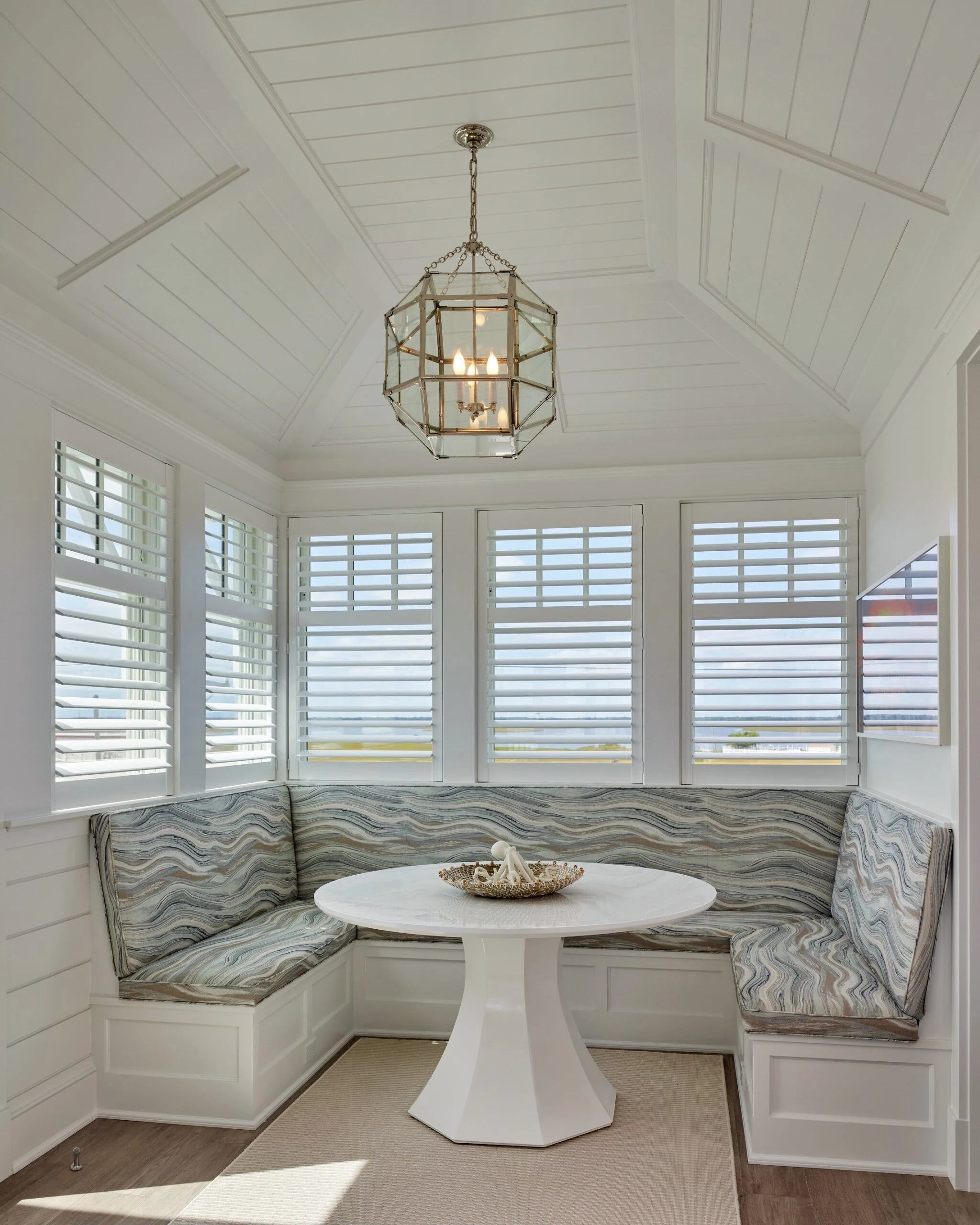 Bright sunroom with vaulted shiplap ceiling, geometric pendant light, built-in banquette seating, pedestal table, and multi-pane shuttered windows.