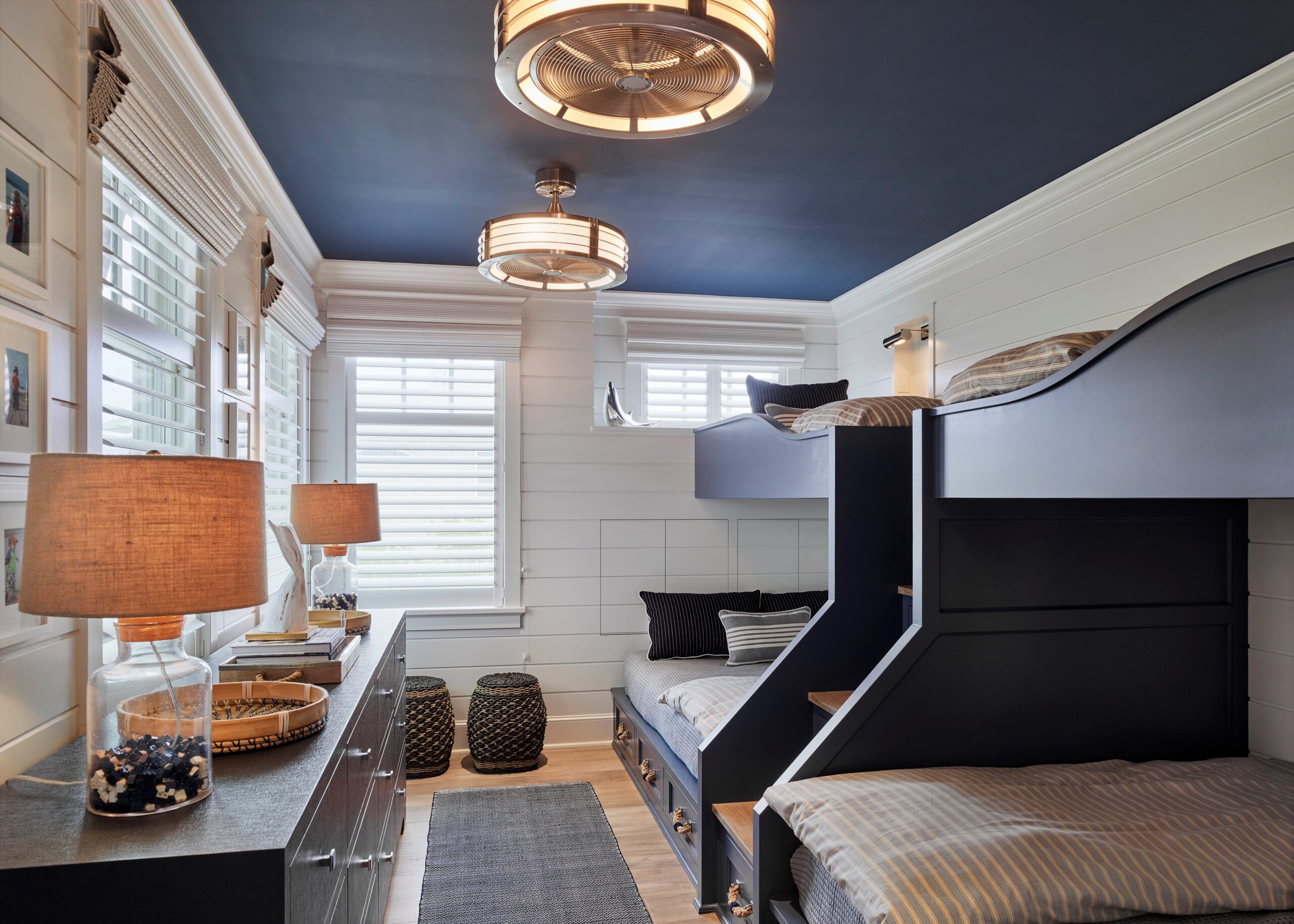 Modern children's bedroom with dual built-in bunk beds, shiplap walls, blue ceiling, brass fixtures, and abundant natural light from shuttered windows.