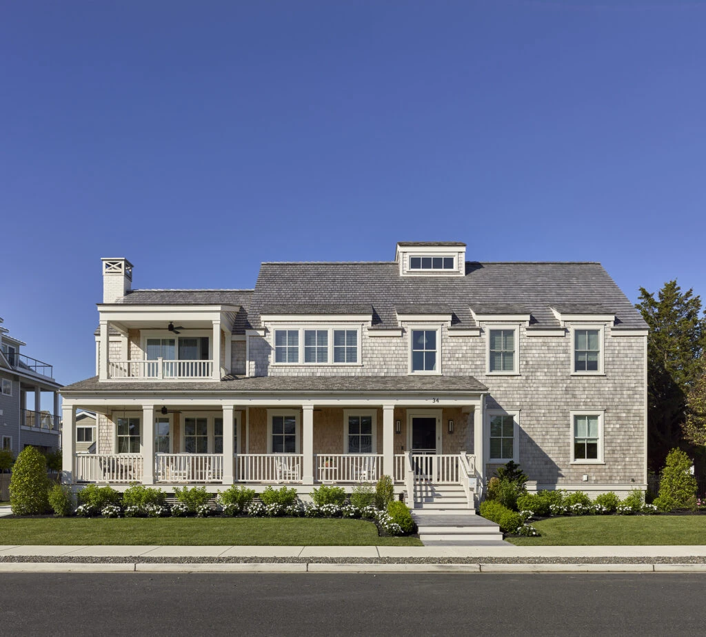 Modern coastal home with shingle facade, wraparound porch, dormer windows, and classical white trim detailing.