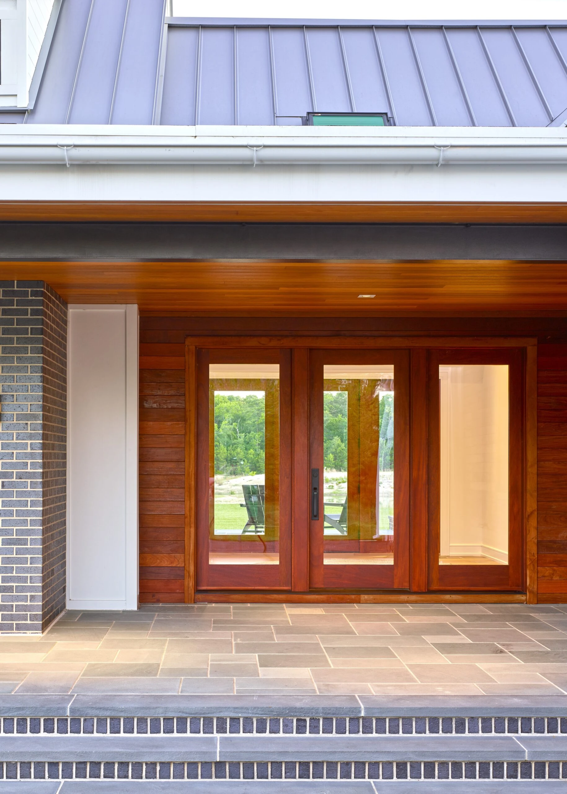 Modern home exterior featuring wood-clad facade, triple glass doors, brick chimney, and standing seam metal roof.