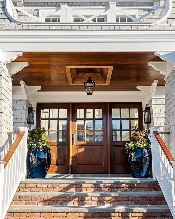 Colonial-style entry with paneled wood doors, white brick facade, exposed wood ceiling, and blue planters flanking the entrance.