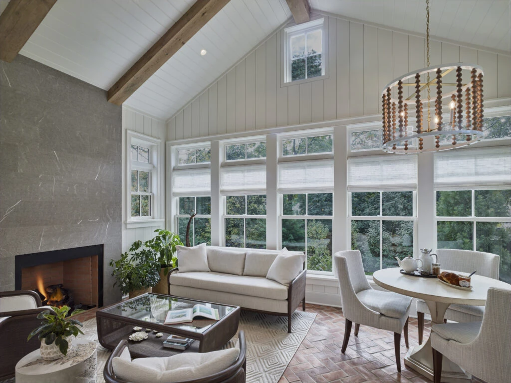 Modern sunroom with vaulted ceiling, exposed beams, extensive glazing, fireplace, and beaded chandelier.