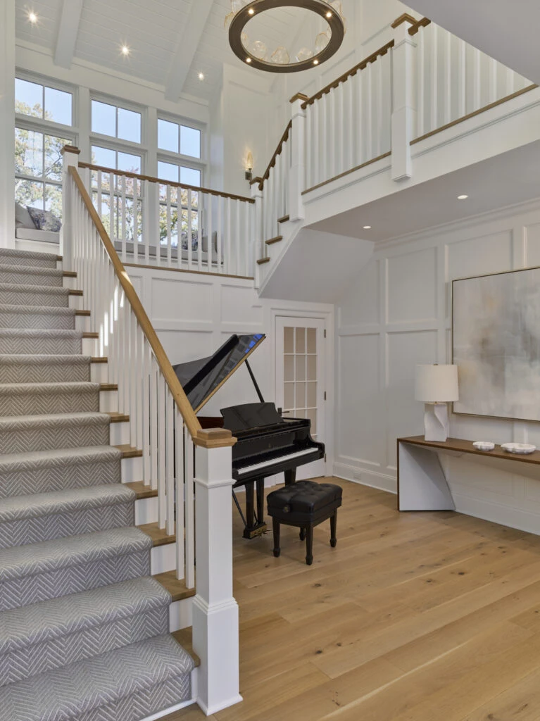 Two-story foyer with grand piano, white shiplap walls, wood handrail, gray chevron stair runner, and clerestory windows.