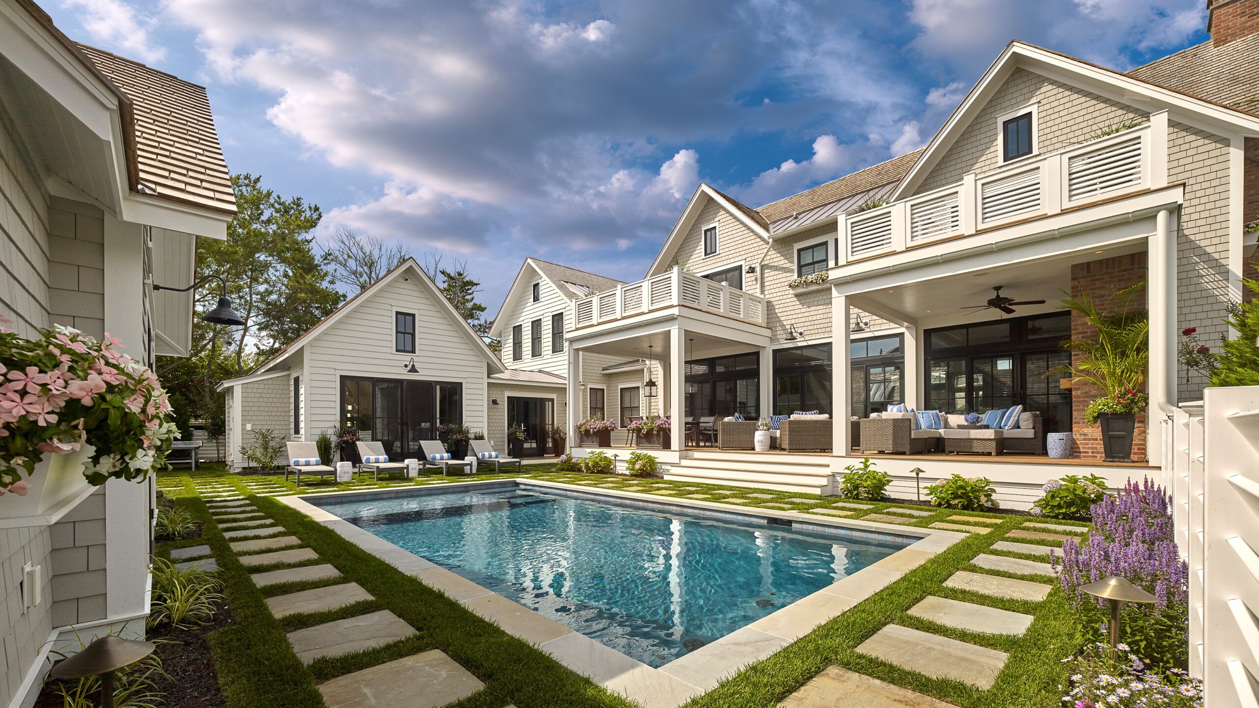 Modern farmhouse with white board-and-batten siding, gable roofs, and covered porches overlooking rectangular pool with stepping stone pathway.