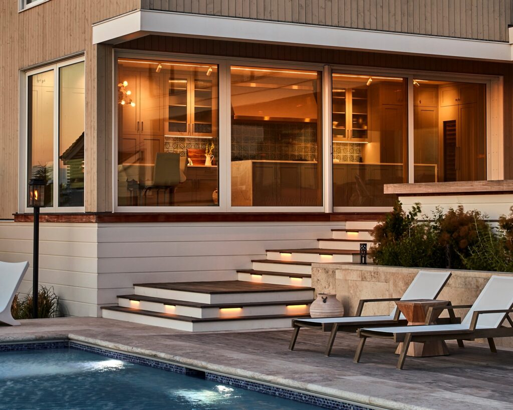 Modern residential elevation with horizontal siding, large glazed doors, integrated step lighting, and poolside terrace at dusk.