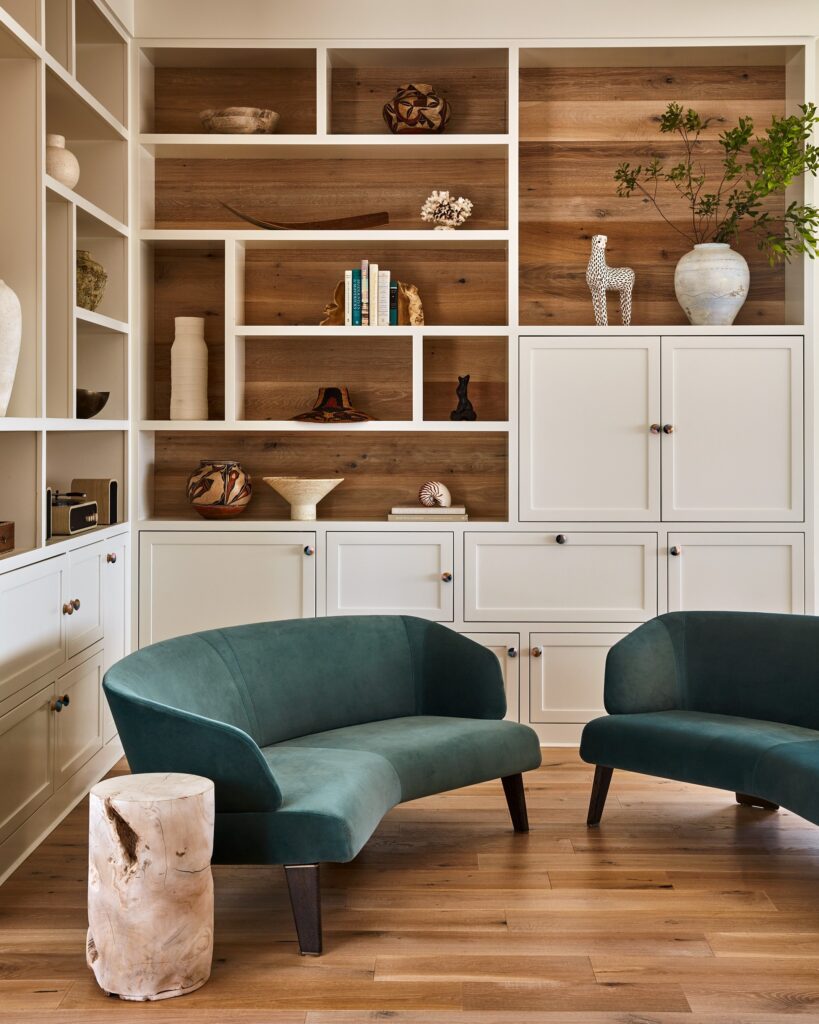Modern living room with built-in shelving unit featuring wooden backing, white cabinetry, and curved teal upholstered seating.
