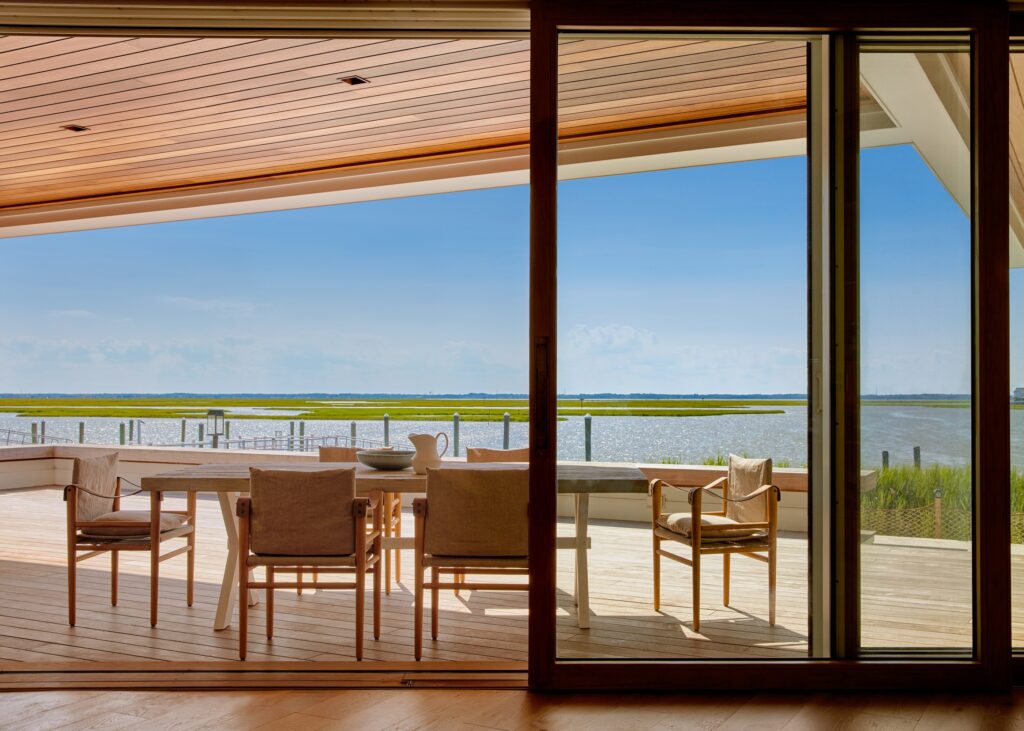 Modern waterfront residence with wood-clad interior, large sliding glass doors, and expansive water views from elevated deck.