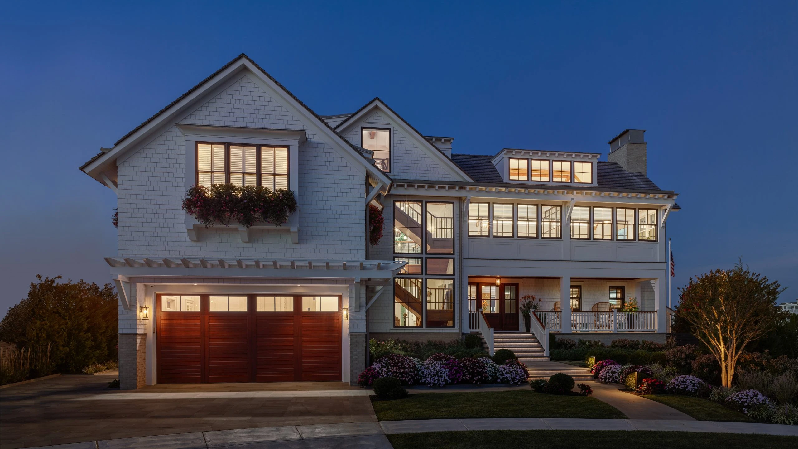 Modern coastal residence with white shingle facade, red garage doors, multi-level design, and extensive glazing at dusk.