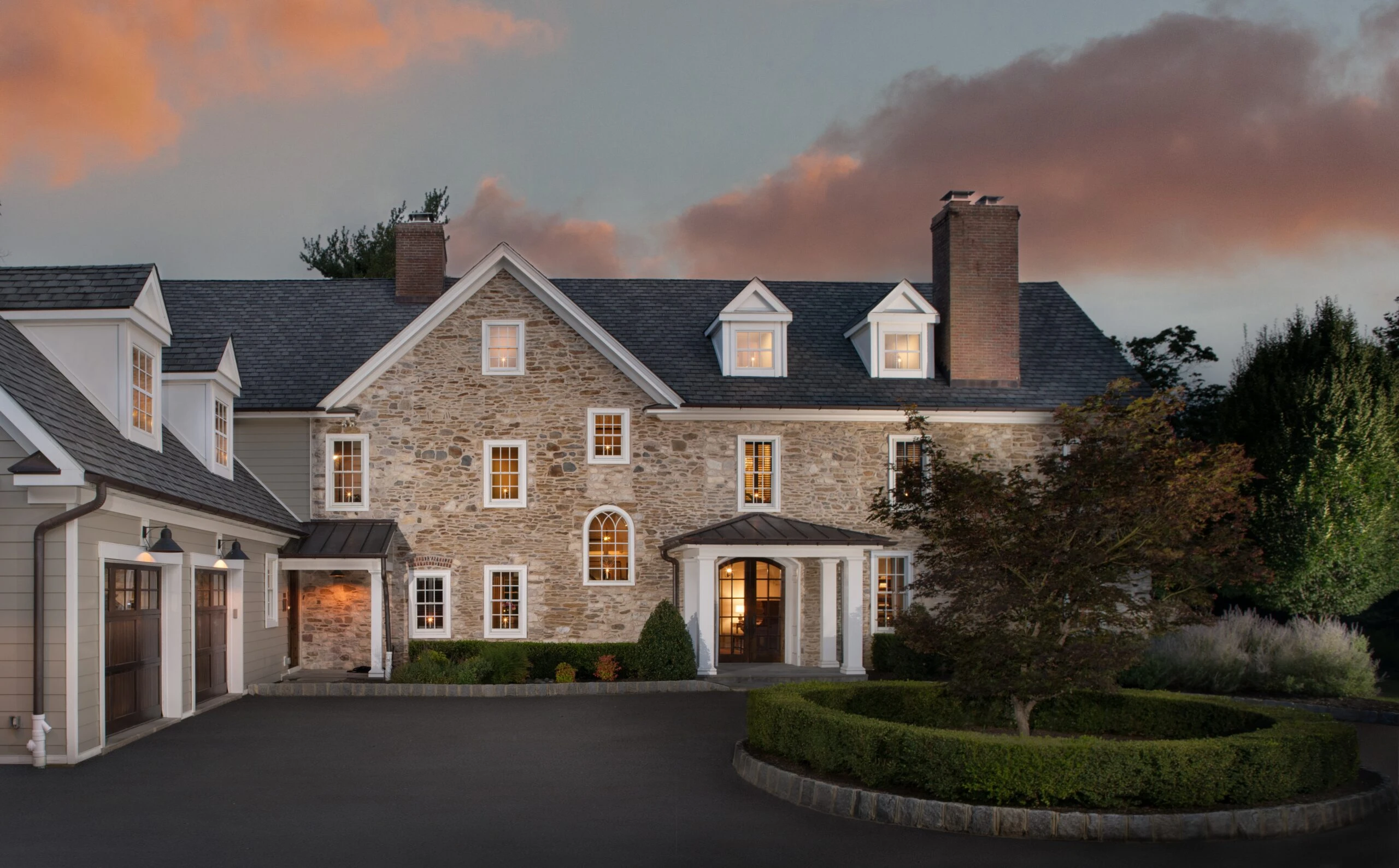 Stone and siding colonial estate with attached garage, arched entry portico, and dormers at dusk.