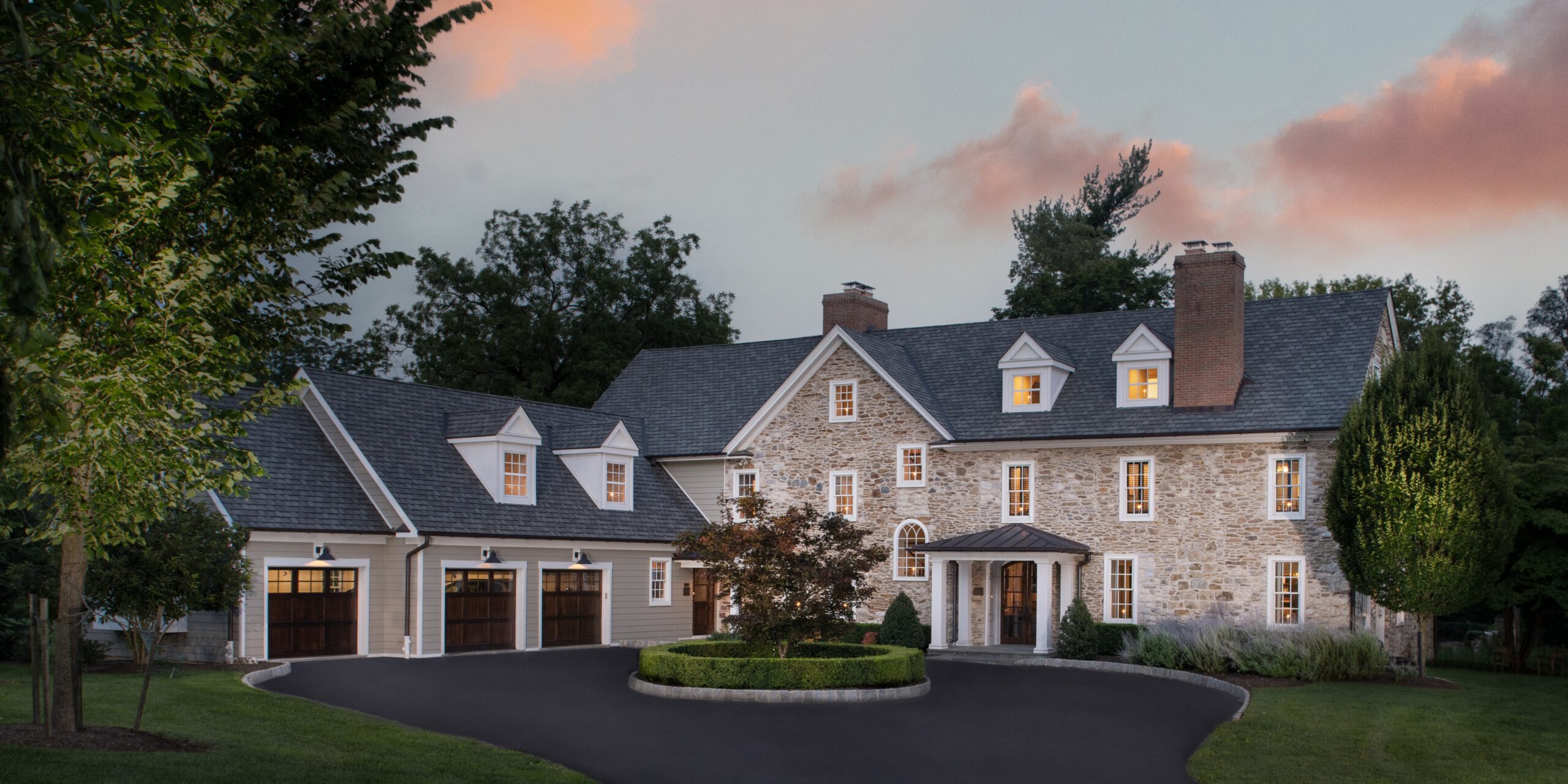 Stone and stucco Colonial Revival estate with three-car garage, dormer windows, and circular driveway at dusk.