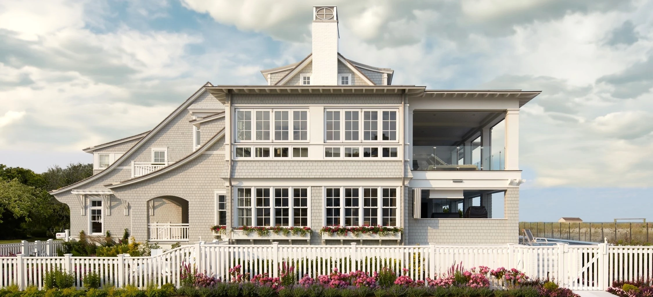 Modern coastal residence with shingle facade, multi-level design, expansive windows, and white picket fence.