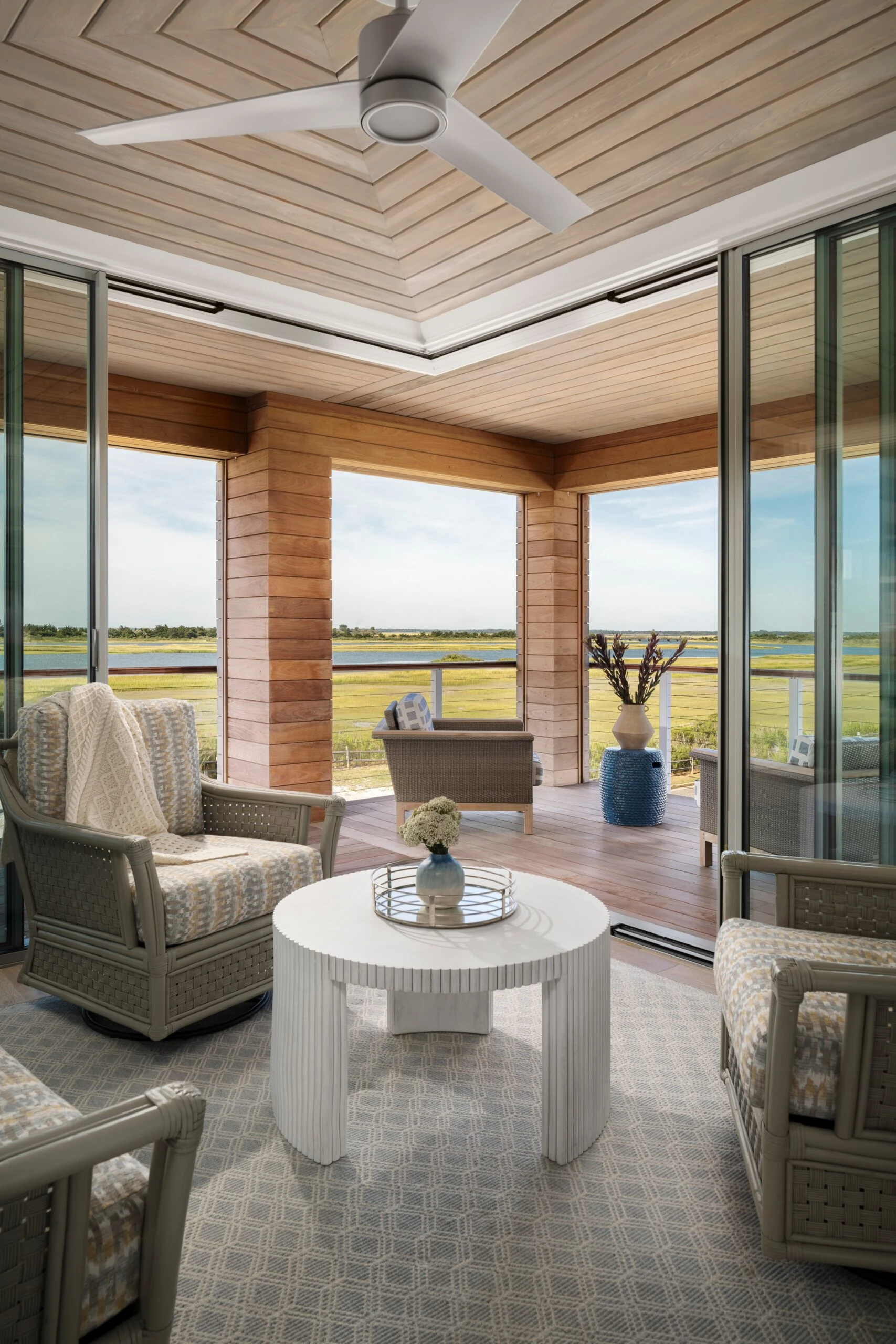 Modern sunroom with coffered wood ceiling, floor-to-ceiling glazing, wicker furnishings, and expansive prairie views.