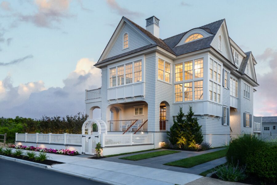 Modern coastal home with blue-gray shingle facade, multi-gabled roofline, extensive glazing, and elevated foundation with arched supports.