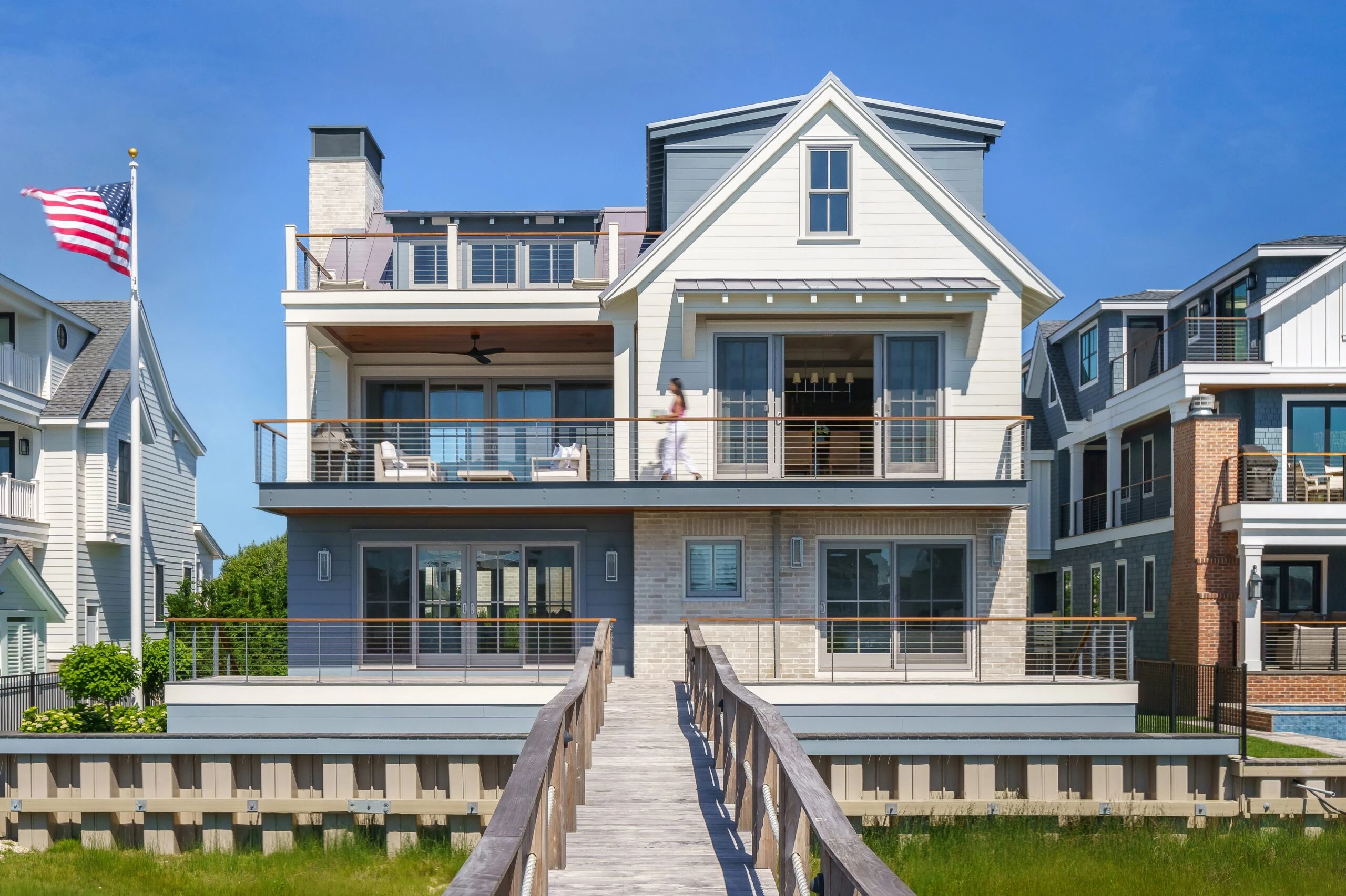 Modern waterfront home with mixed materials: white siding upper floors, stone and blue-grey lower level, horizontal railings, and gabled roofline.