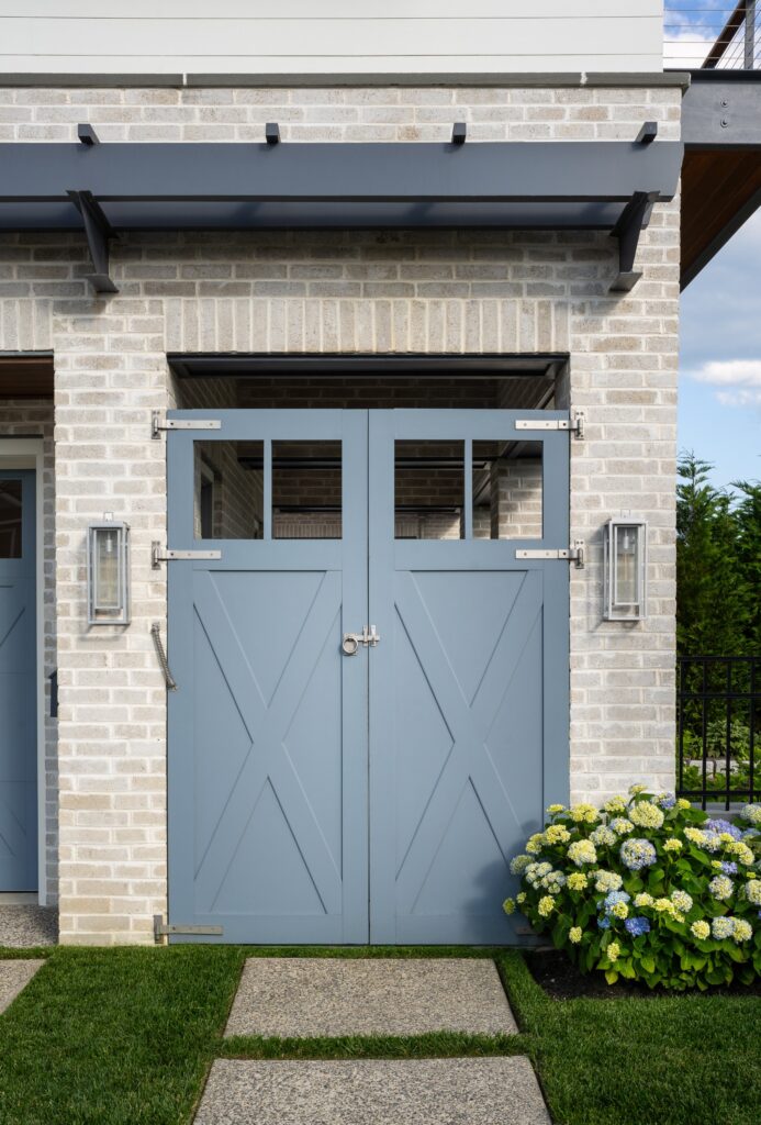 Light blue carriage doors with X-pattern panels on brick facade, black steel pergola overhead, and flanking sconces.