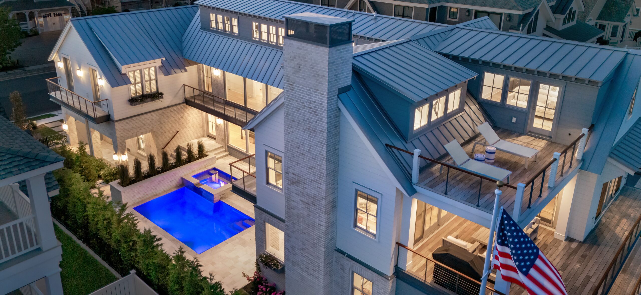 Modern residential home with metal roof, brick chimney, illuminated windows, and blue-lit rectangular pool on paved terrace.