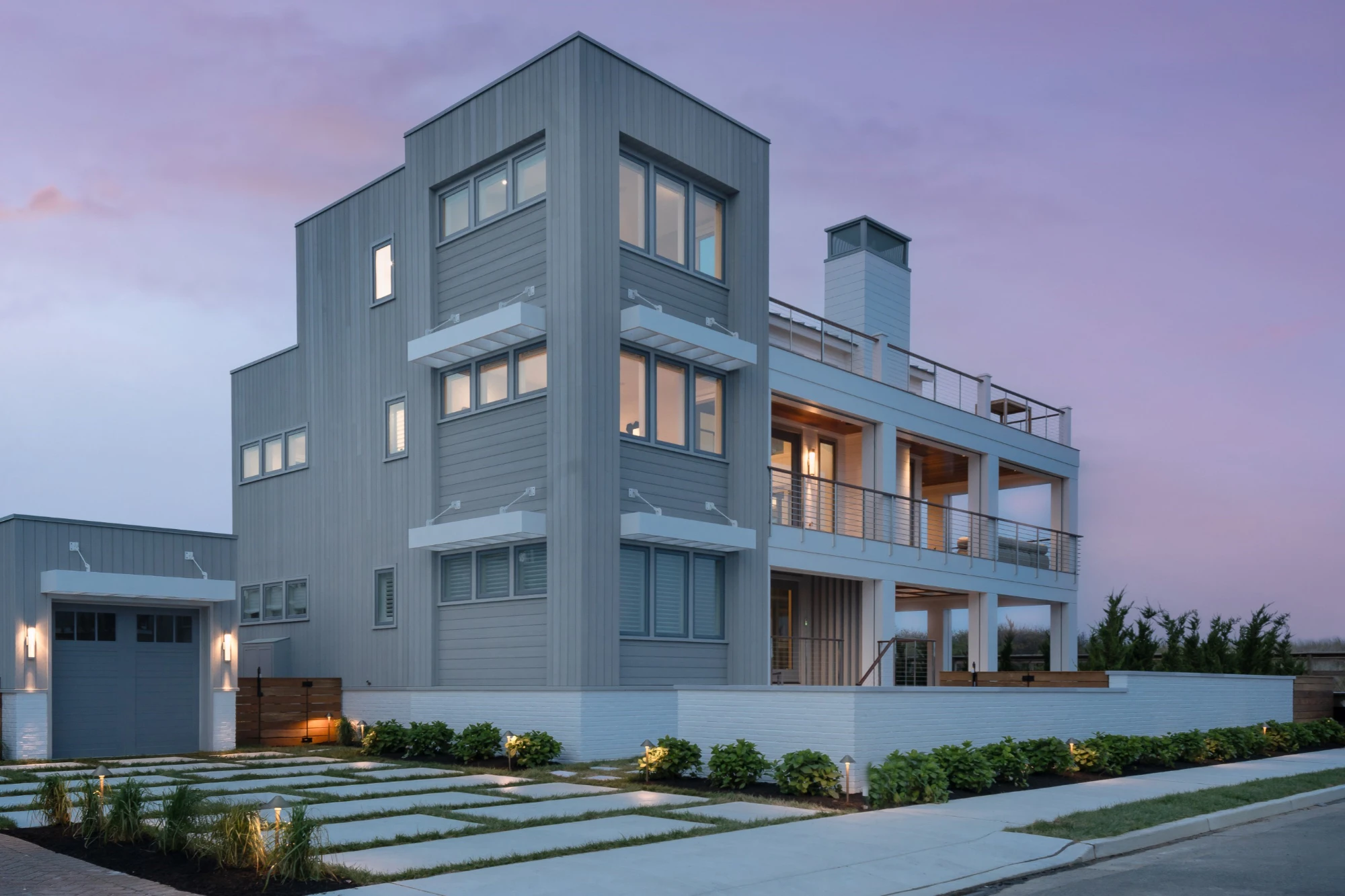 Modern coastal residence with vertical metal siding, stacked windows, open deck balconies, and white base foundation at dusk.