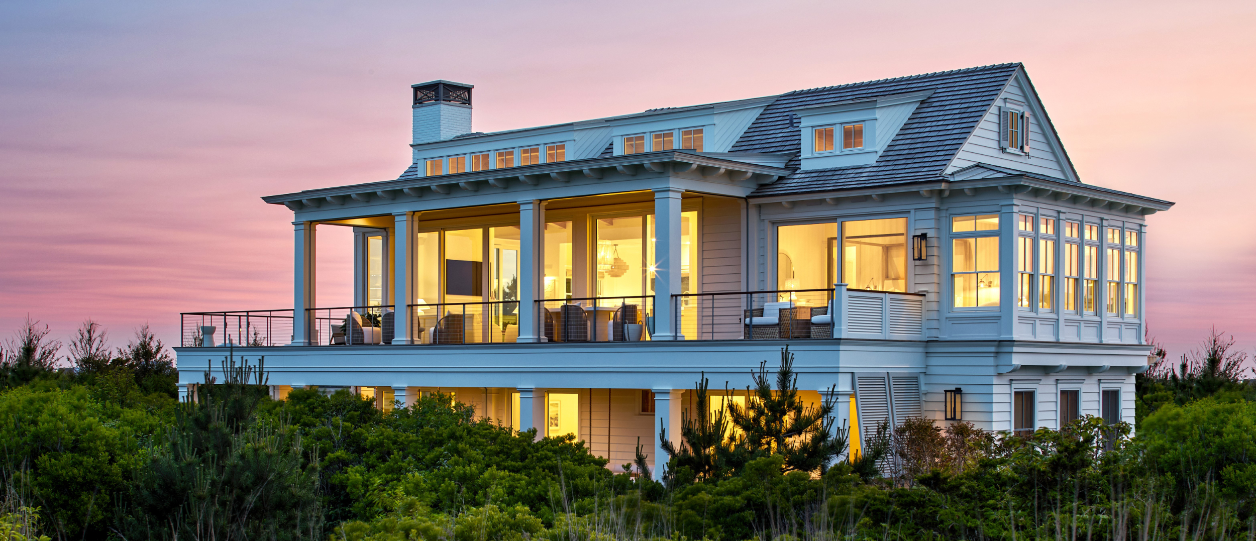 Modern coastal residence with elevated first floor, wraparound deck, metal roof, and extensive glazing at dusk.
